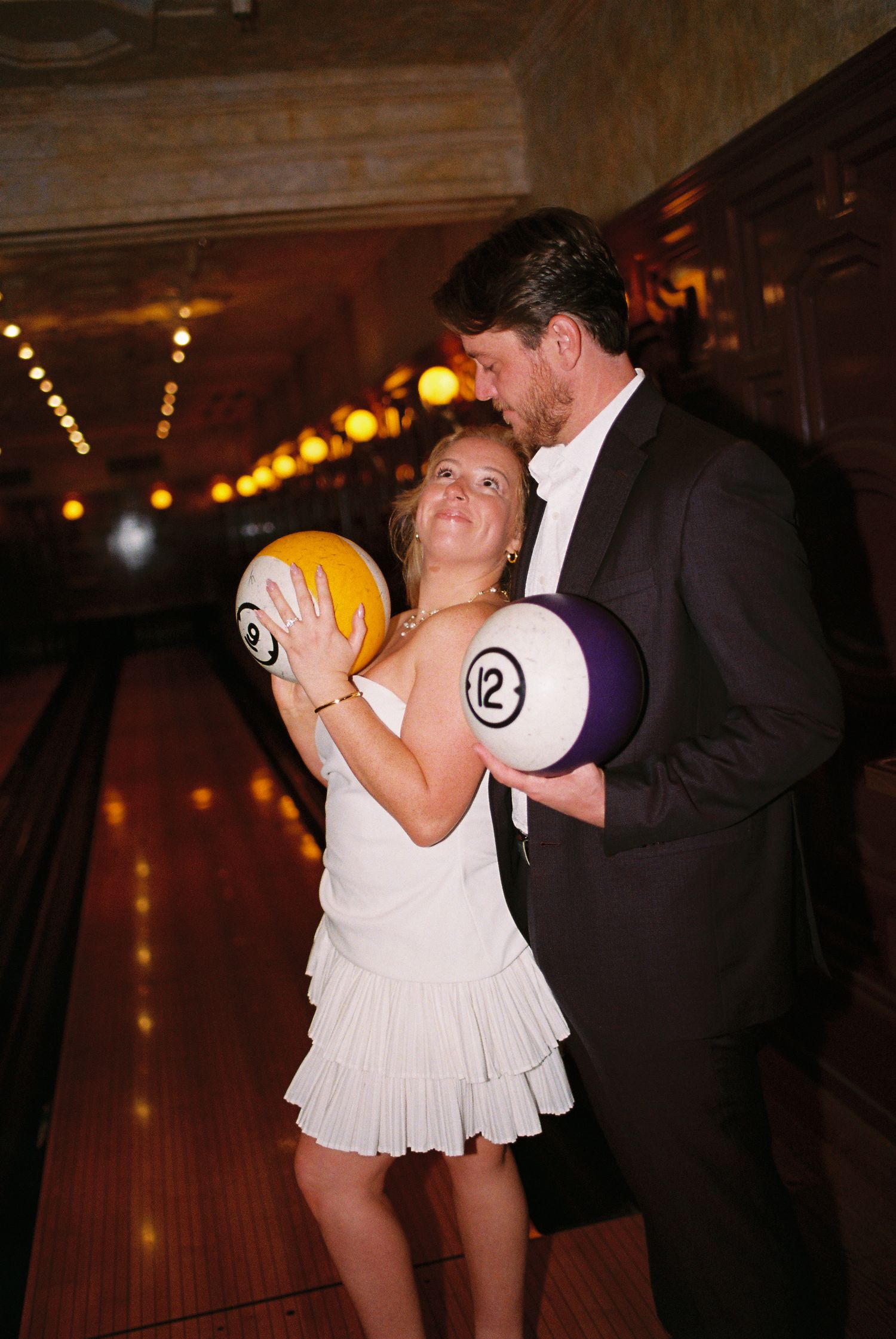 Couple in formal attire holding oversized billiard balls at a bowling alley, perfect for a unique wedding photo.