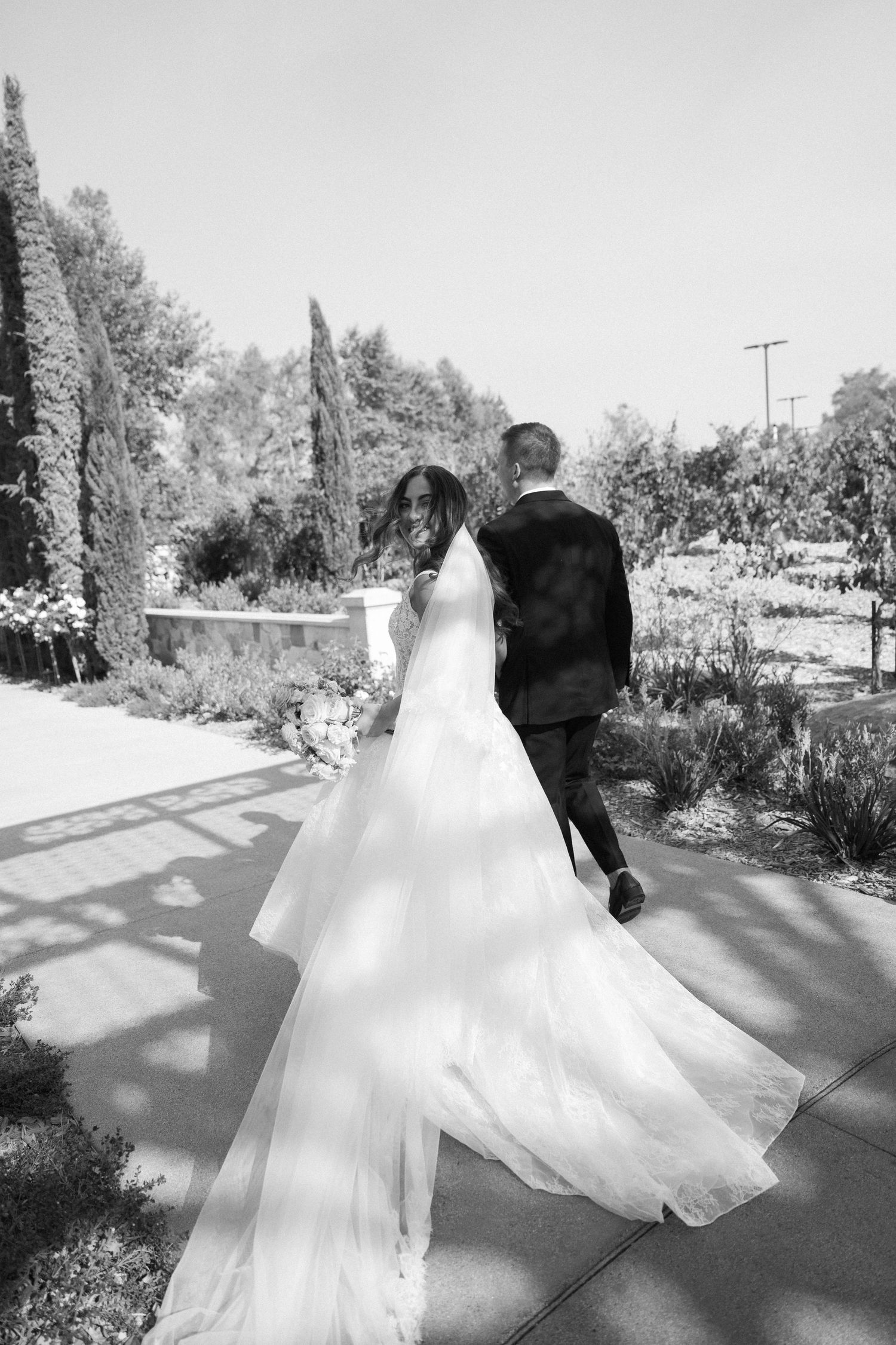 Black and white photo of bride and groom walking hand in hand through a garden, bride's gown flowing behind her.