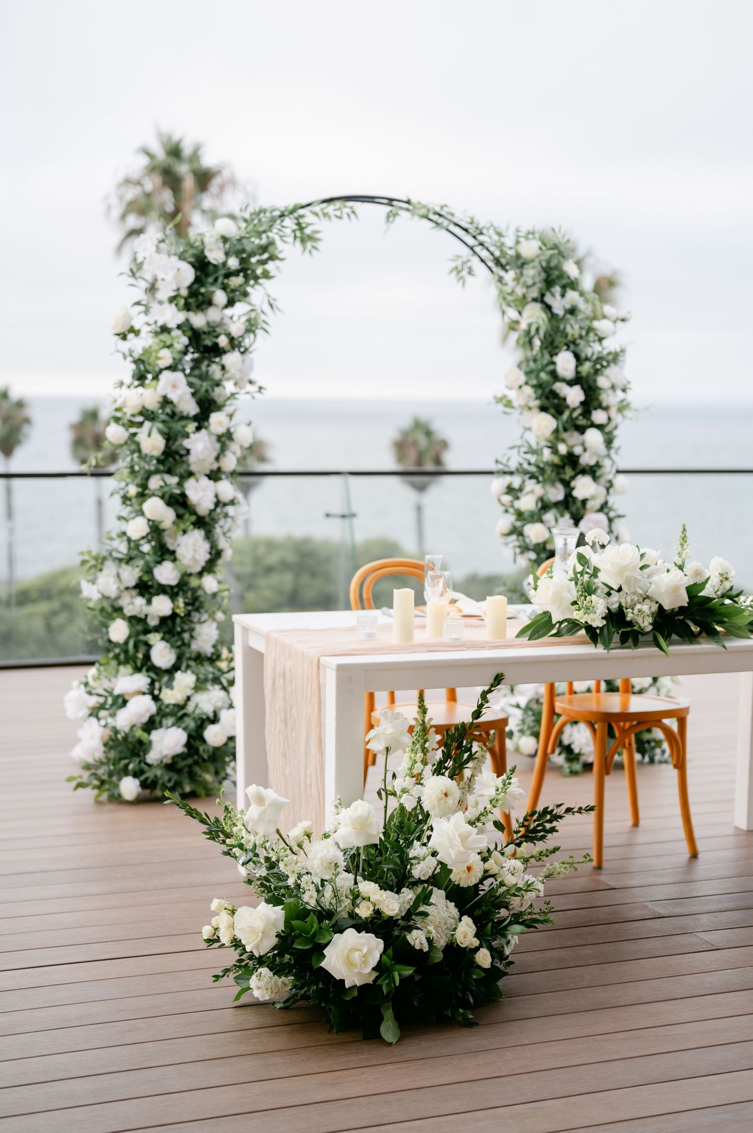 White floral arch arrangements frame wooden chairs at outdoor wedding ceremony setup.