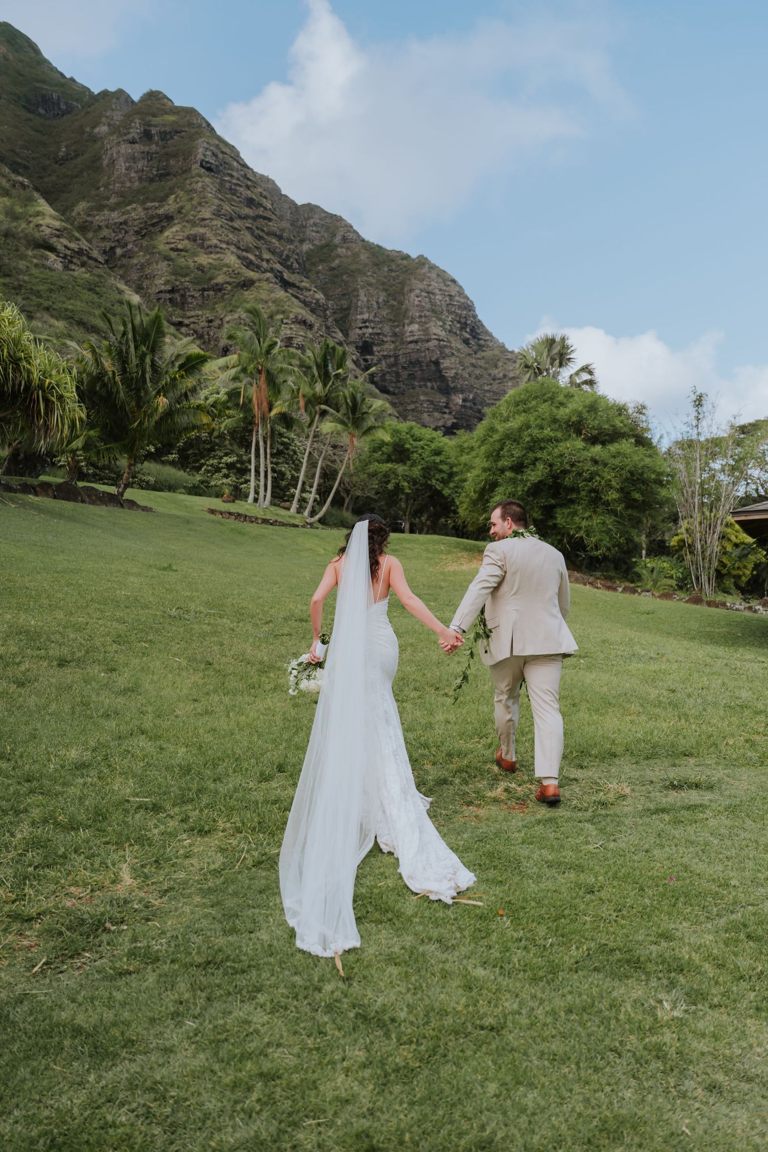 Two people in white outfits walk together across a lush green lawn with dramatic mountain cliffs in the background.