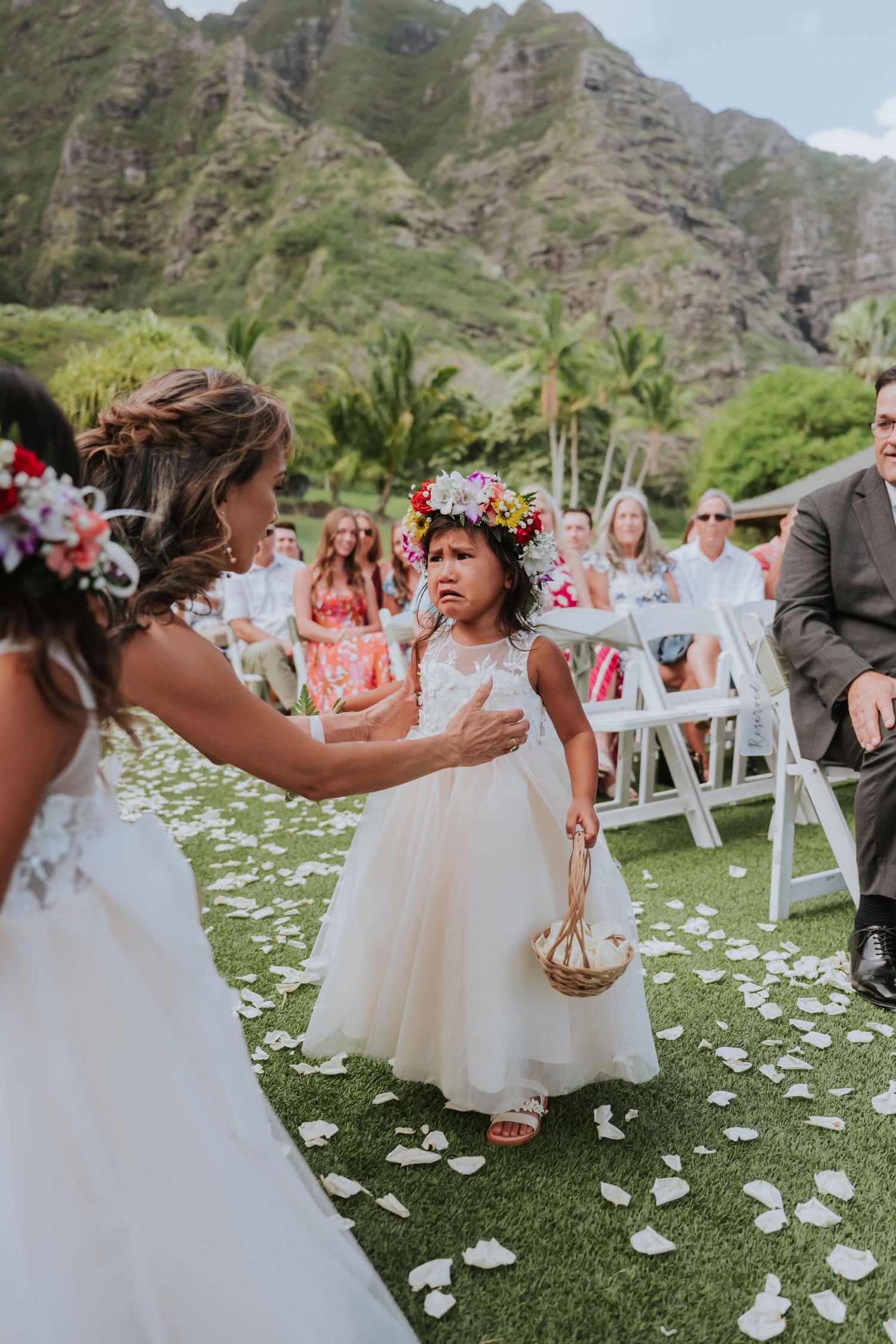 Flower girls in white dresses walking down aisle scattered with petals against mountain backdrop.