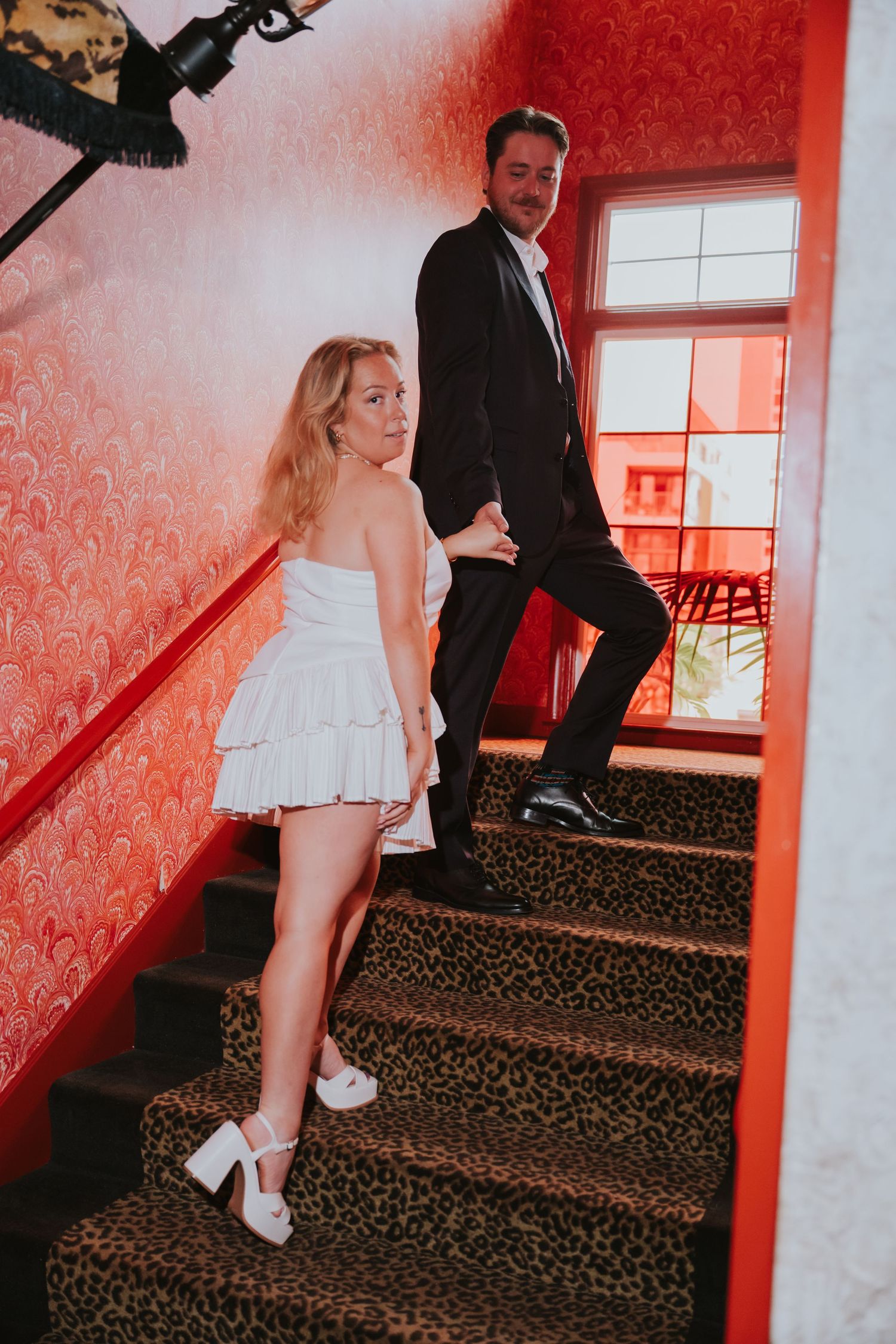Bride in white mini dress and groom in black tuxedo descend leopard-print stairs in a red wallpapered venue.