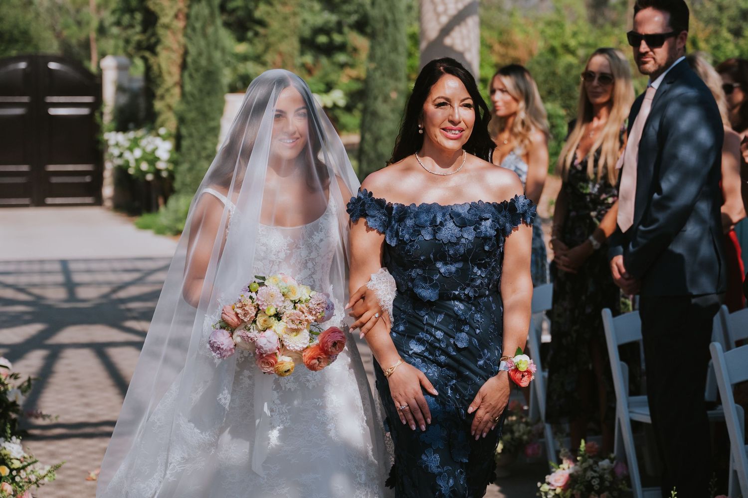 Bride in white gown and veil walks down outdoor aisle with woman in blue floral dress at sunny wedding ceremony.