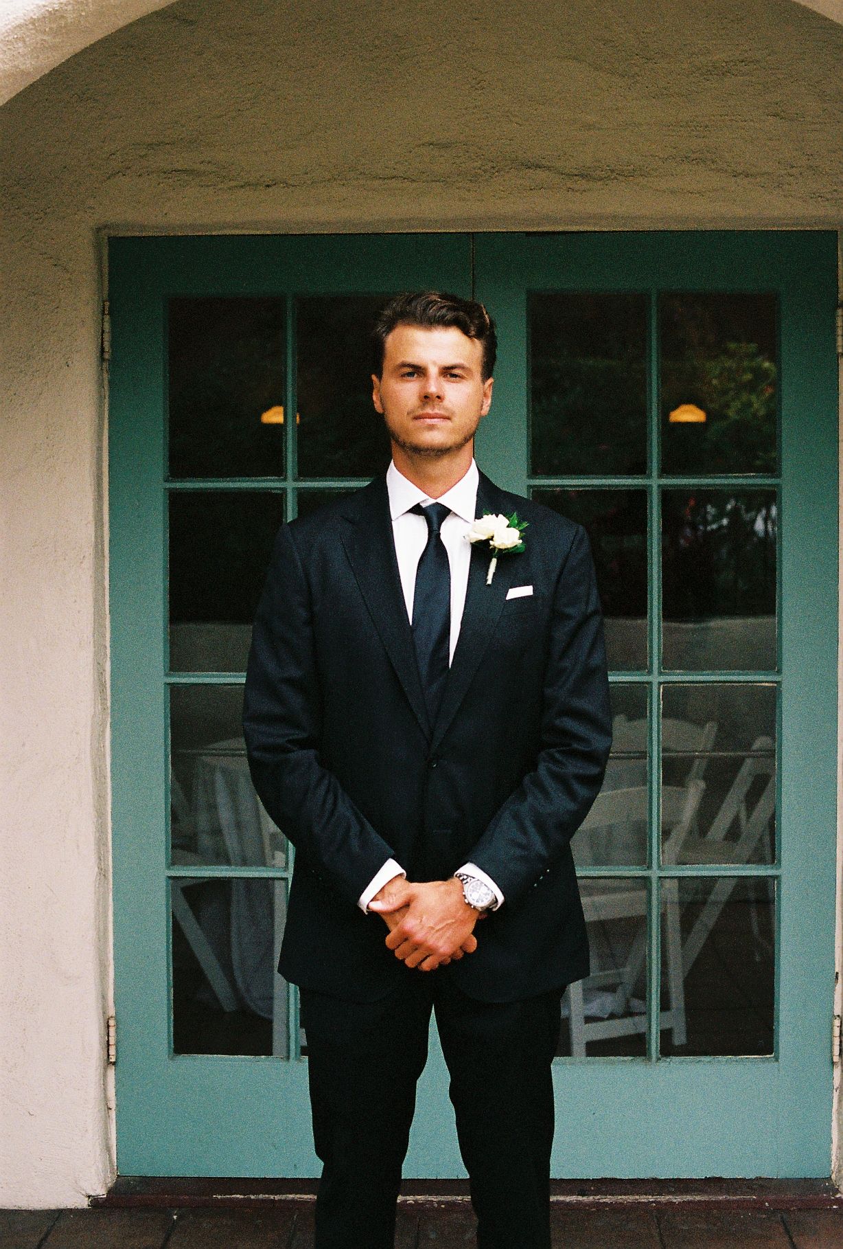 Groom in dark navy suit with white boutonniere and black tie standing before teal French doors.