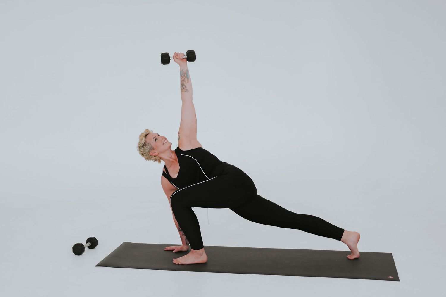 Person demonstrates side plank with dumbbell raise exercise on yoga mat in studio setting.