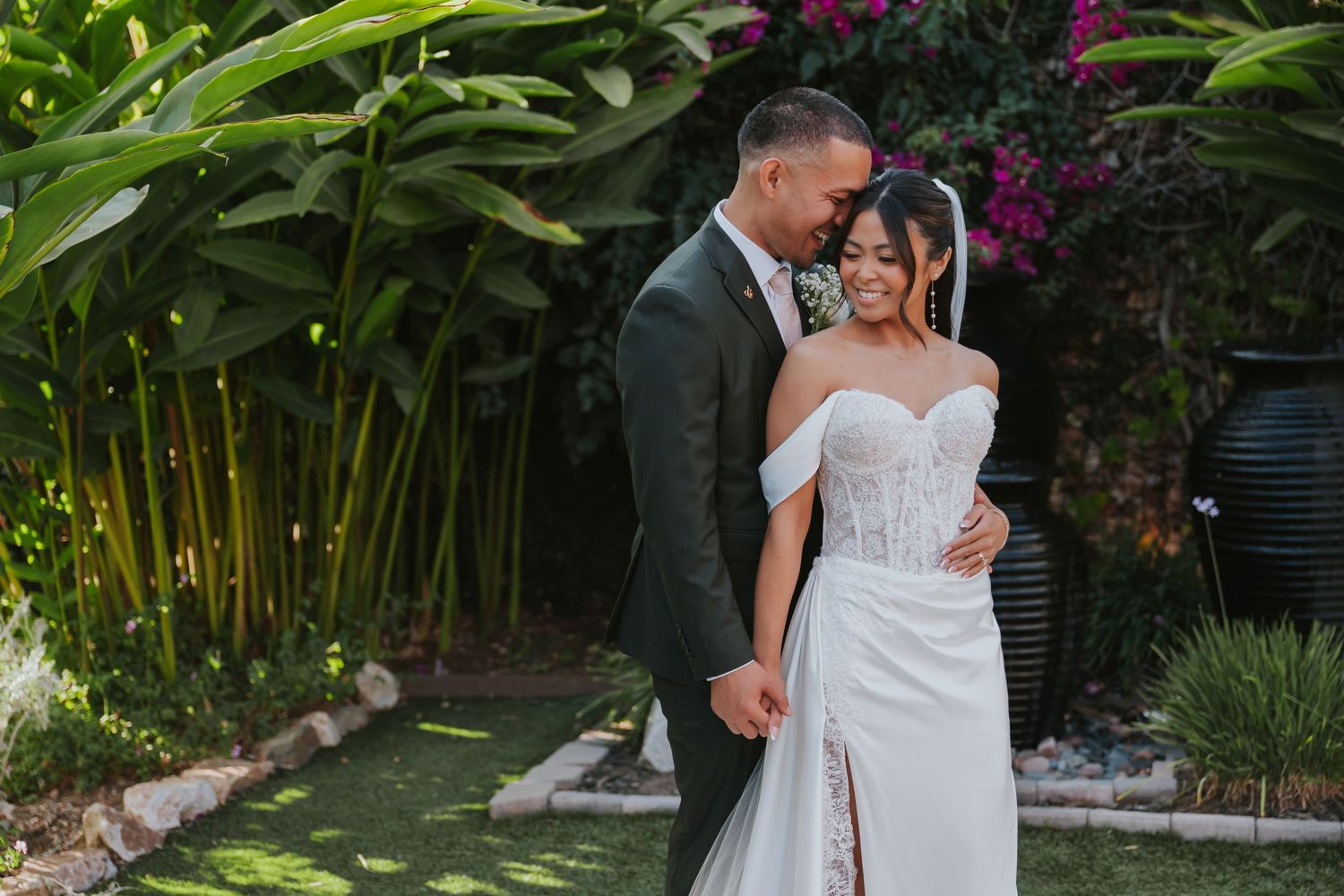 Wedding couple poses in a lush garden setting with tropical plants and pink flowers in the background.