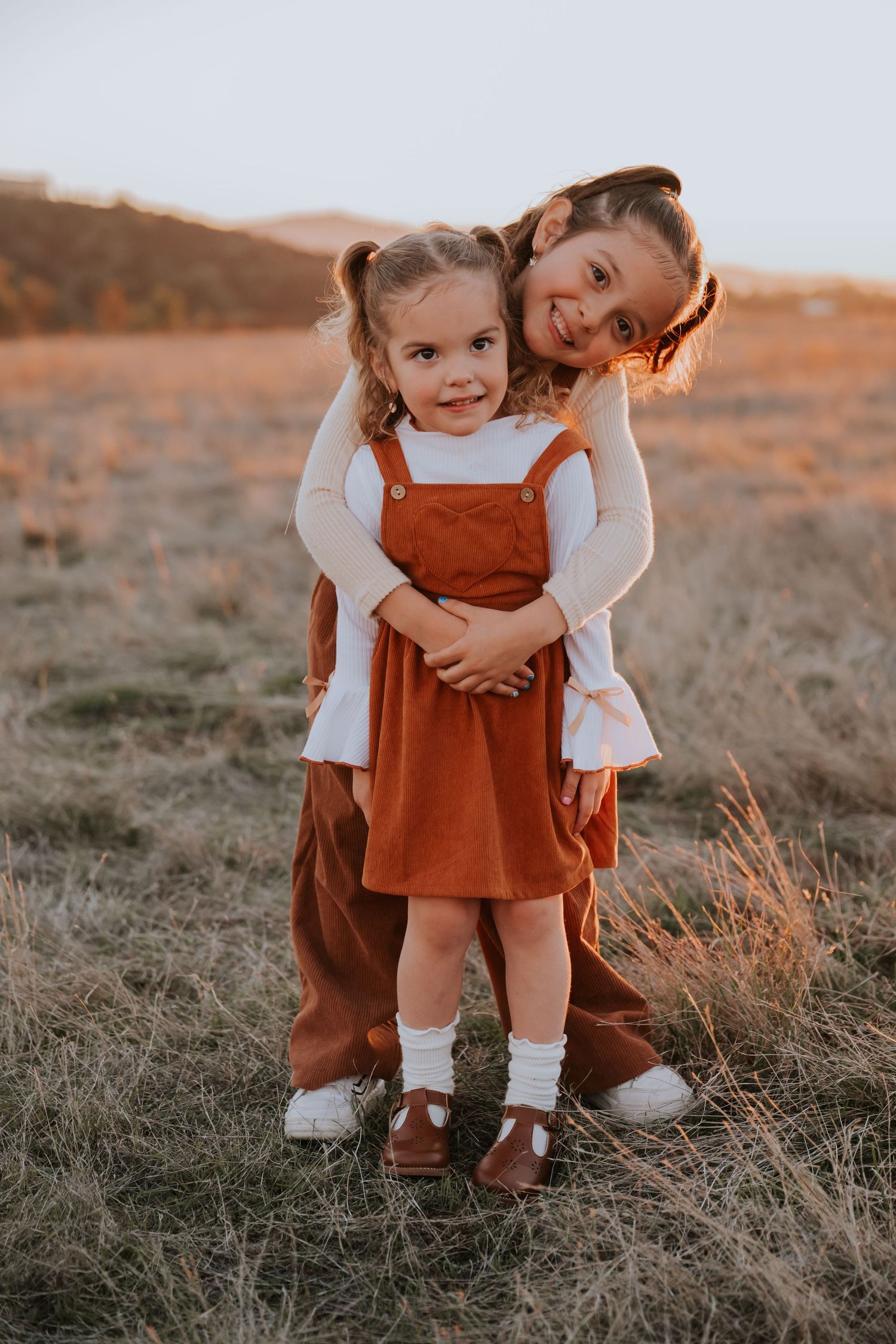 Two young sisters hugging in a golden field at sunset, wearing rust-colored outfits and white shirts.