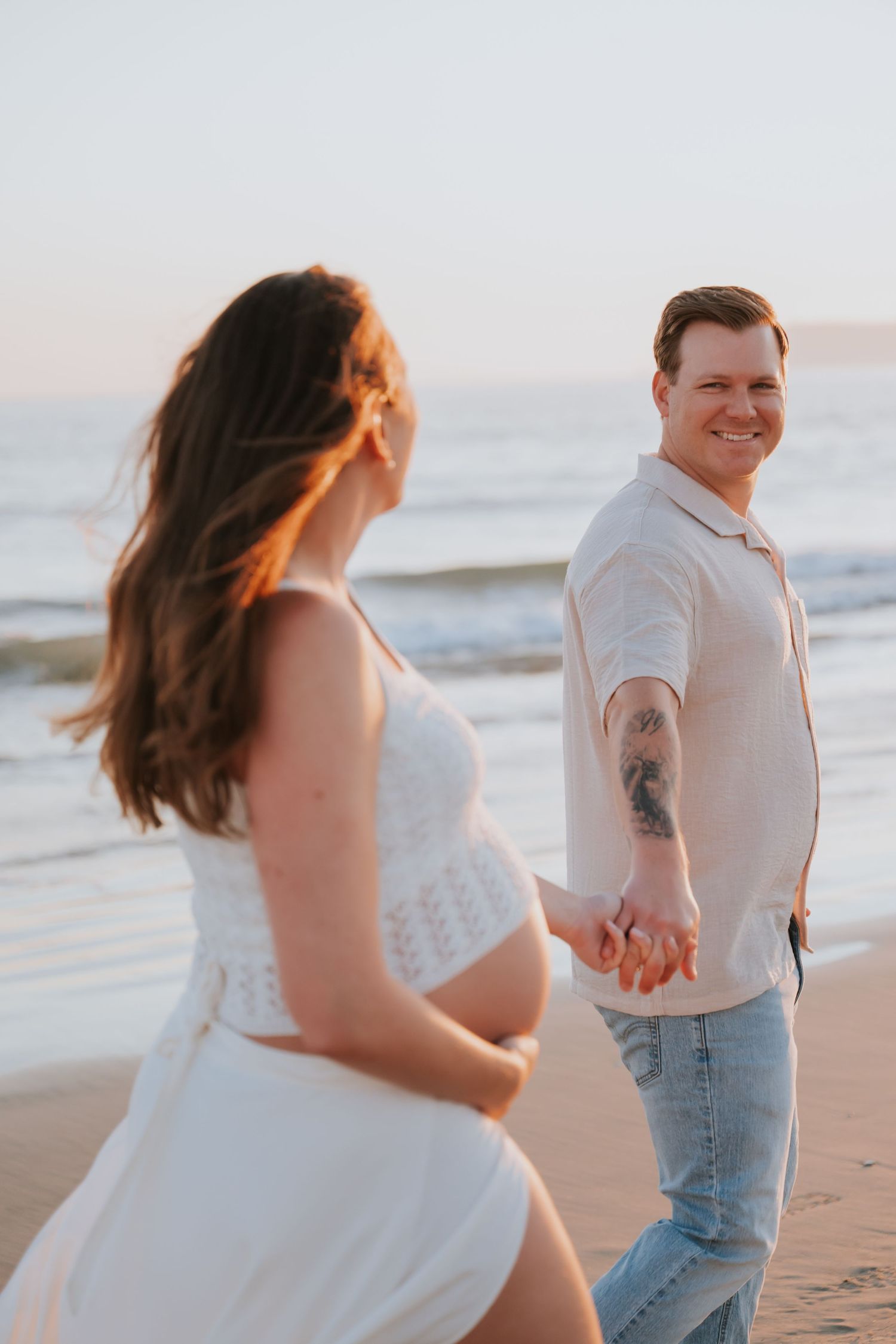 Pregnant couple holding hands on a beach at sunset, man smiling back at camera wearing white shirt and jeans.