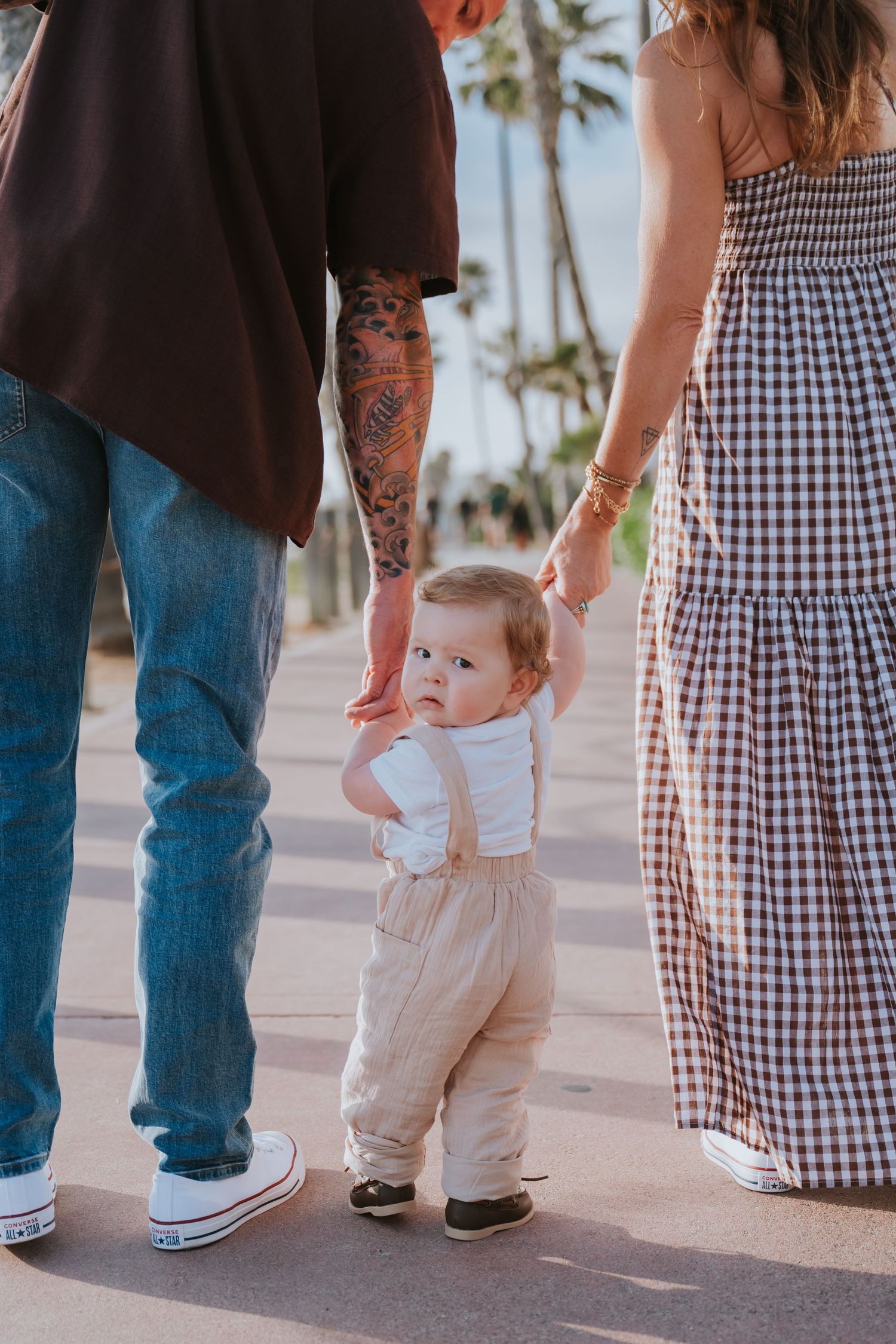 A baby in suspenders walks between parents, one with tattoos, on a palm-lined path.