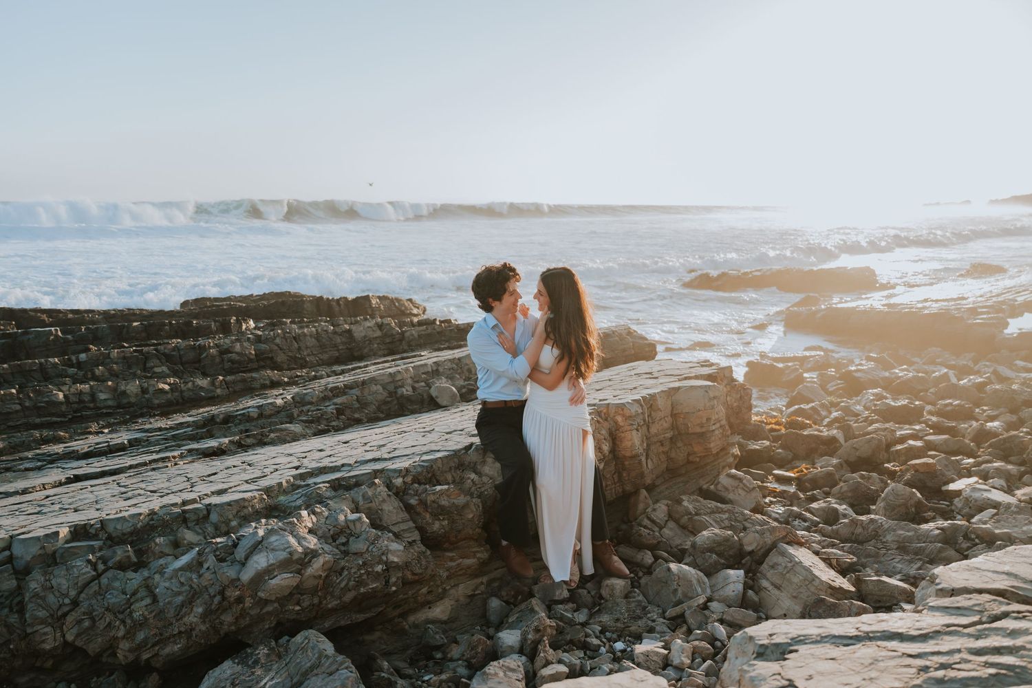 Couple embracing on rocky coastal cliffs at golden hour with ocean waves in the background.