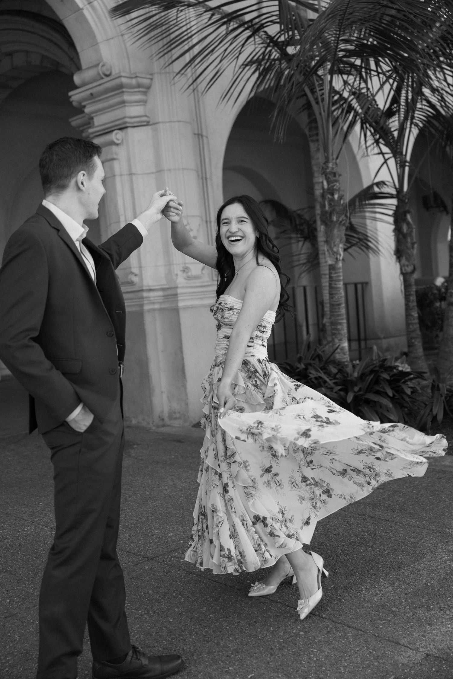 Black and white photo of a couple dancing outdoors, woman in floral dress twirling, man in suit, arched building backdrop.