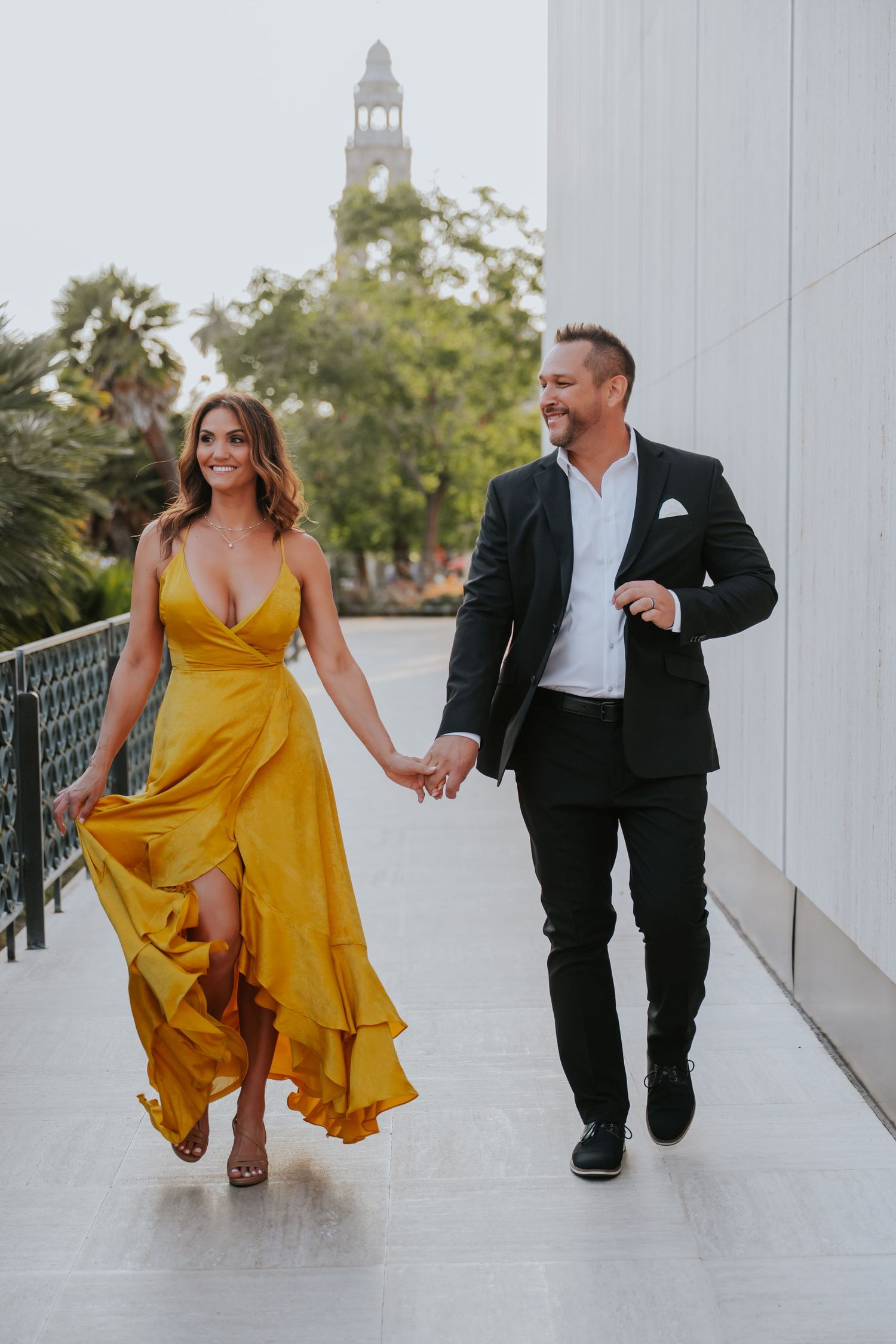 Couple walking hand-in-hand, woman in yellow dress, man in black suit, with a tower and palm trees in the background.
