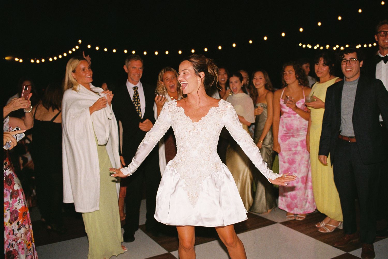 Bride in short white lace long-sleeve wedding dress dancing joyfully at outdoor reception with string lights.