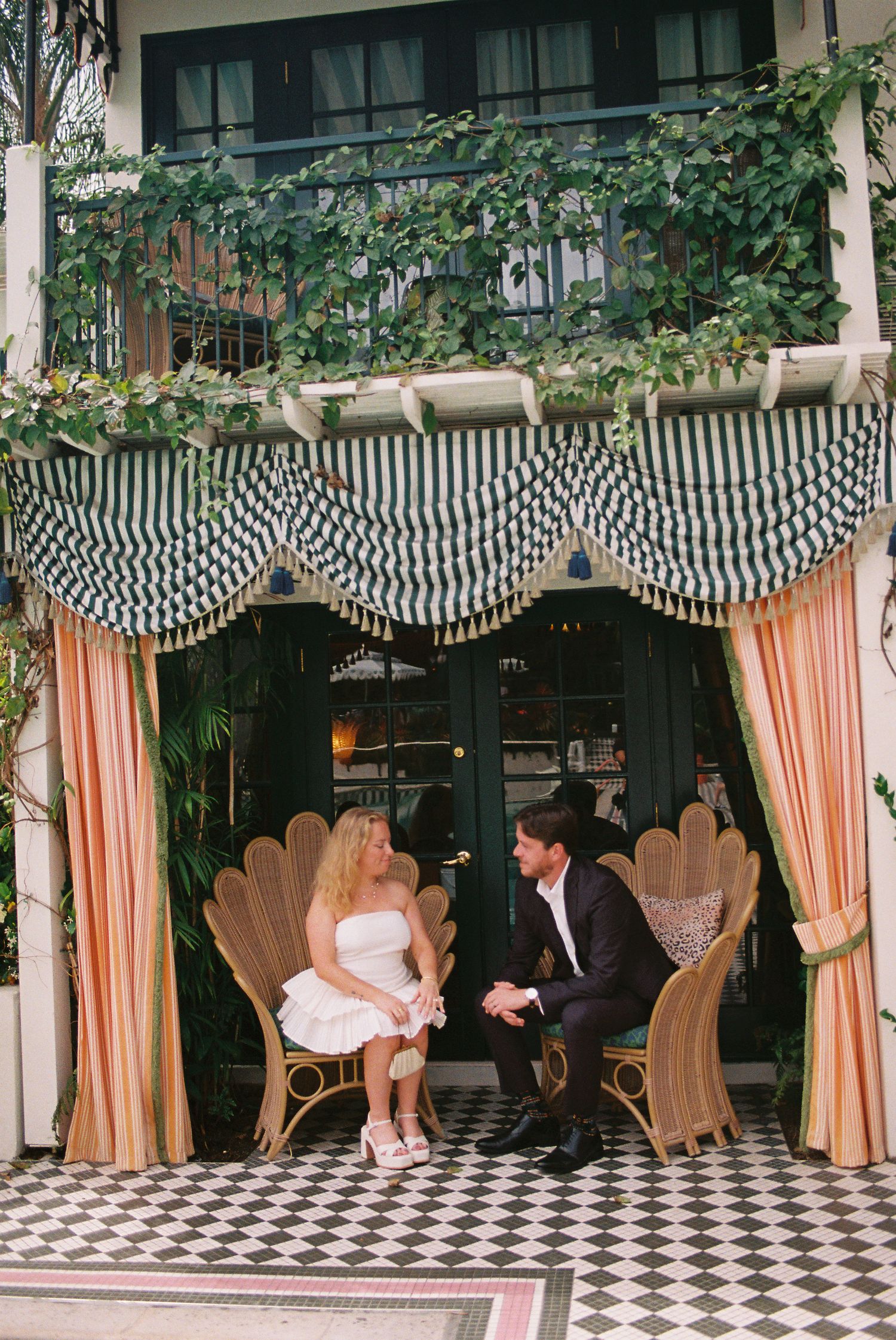 Couple sitting in wicker peacock chairs under striped canopy with pink curtains at elegant venue.