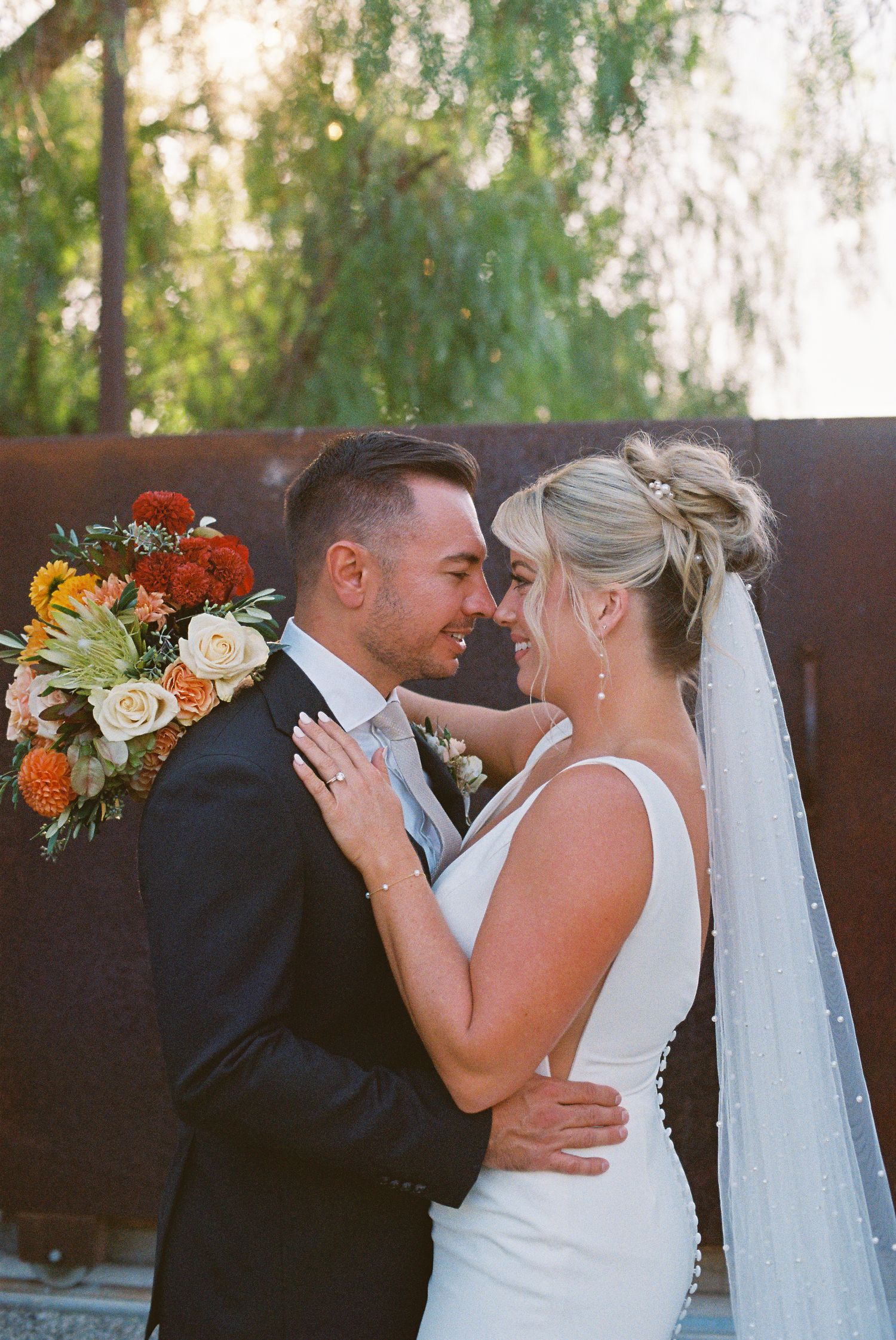 Bride and groom share a romantic moment, she holds a vibrant autumn bouquet, wearing a white gown and veil outdoors.