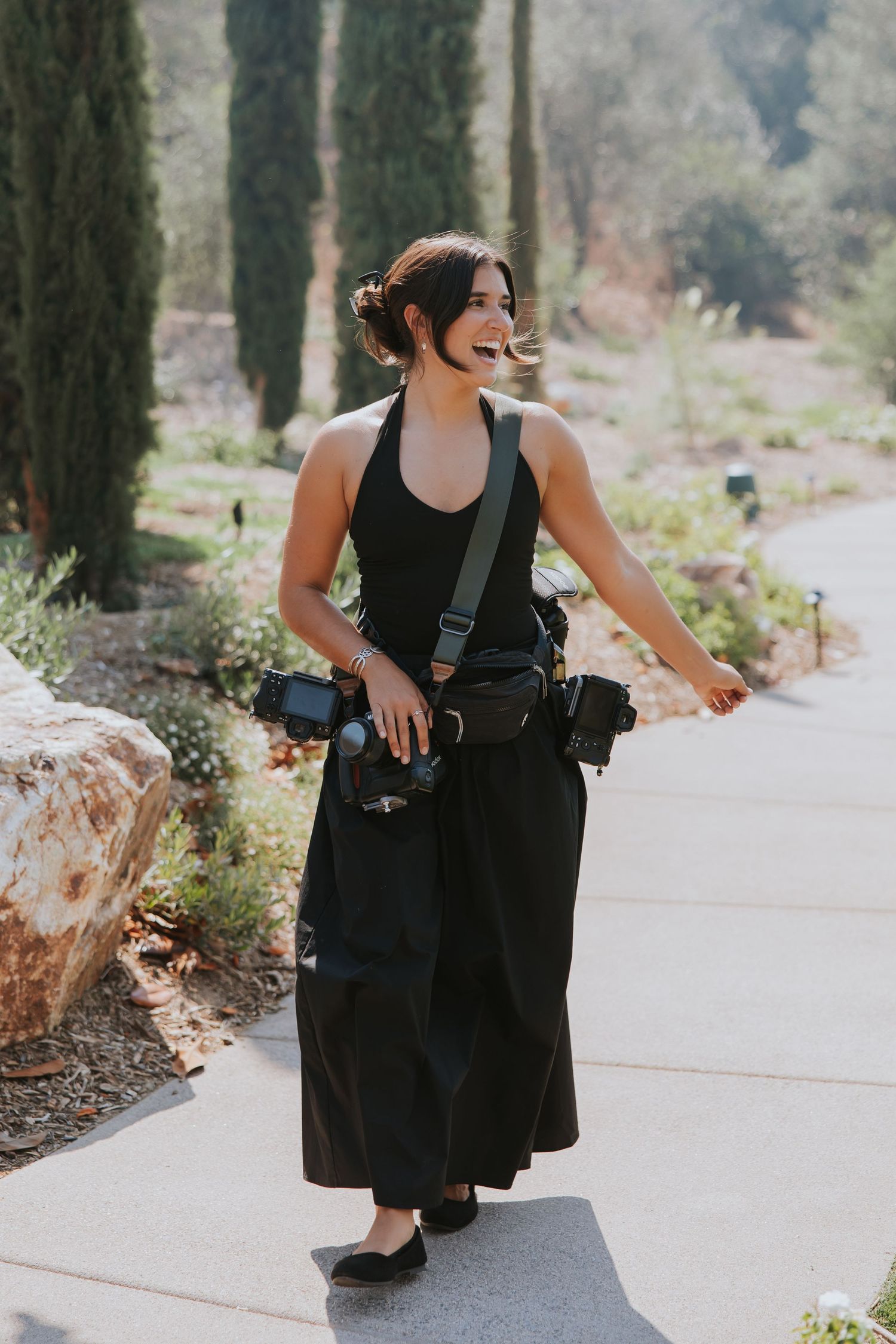 A smiling female photographer in all-black outfit carrying camera gear walks along a garden pathway.