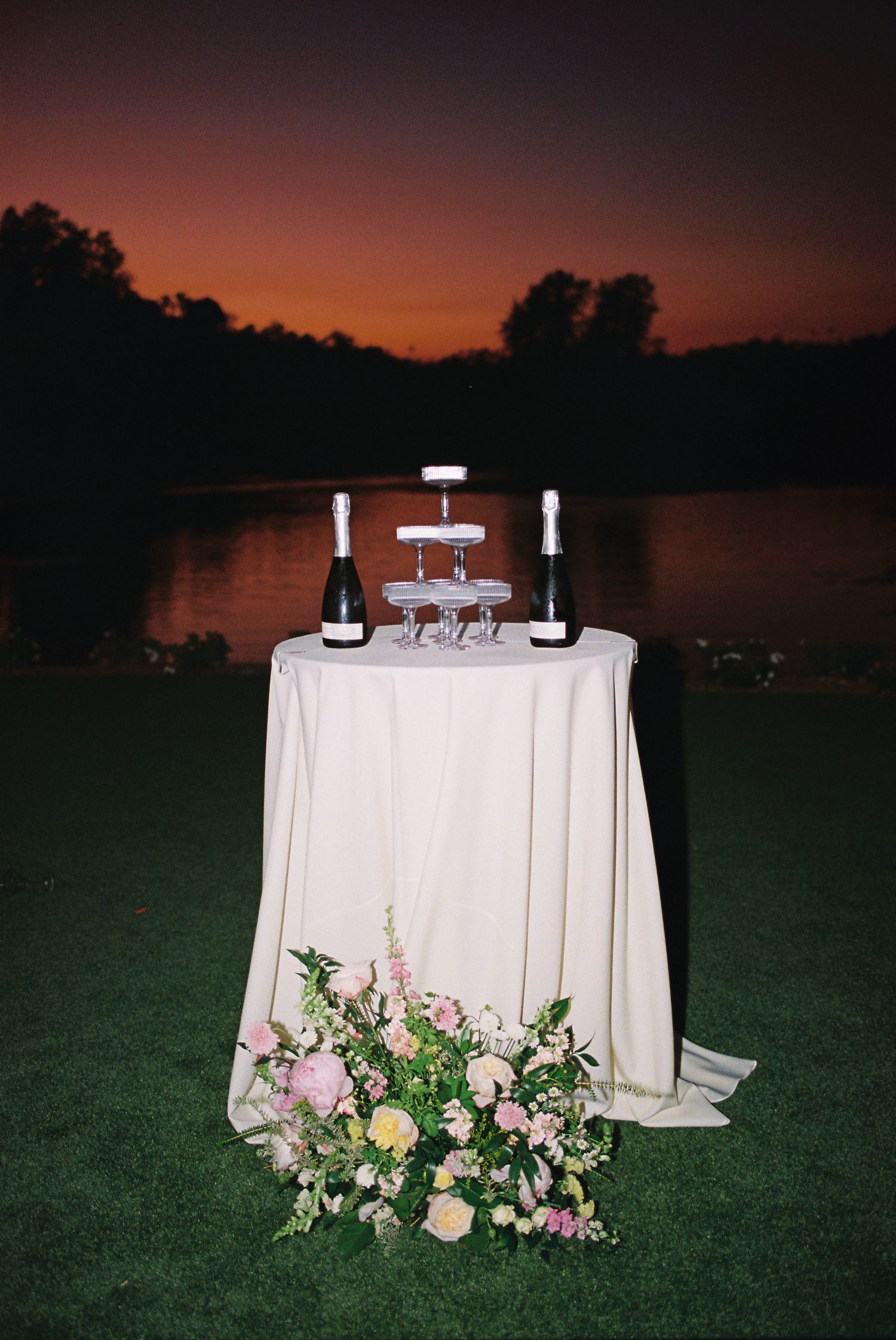 Elegant champagne tower on a white draped table with floral arrangement at sunset by a serene lake.