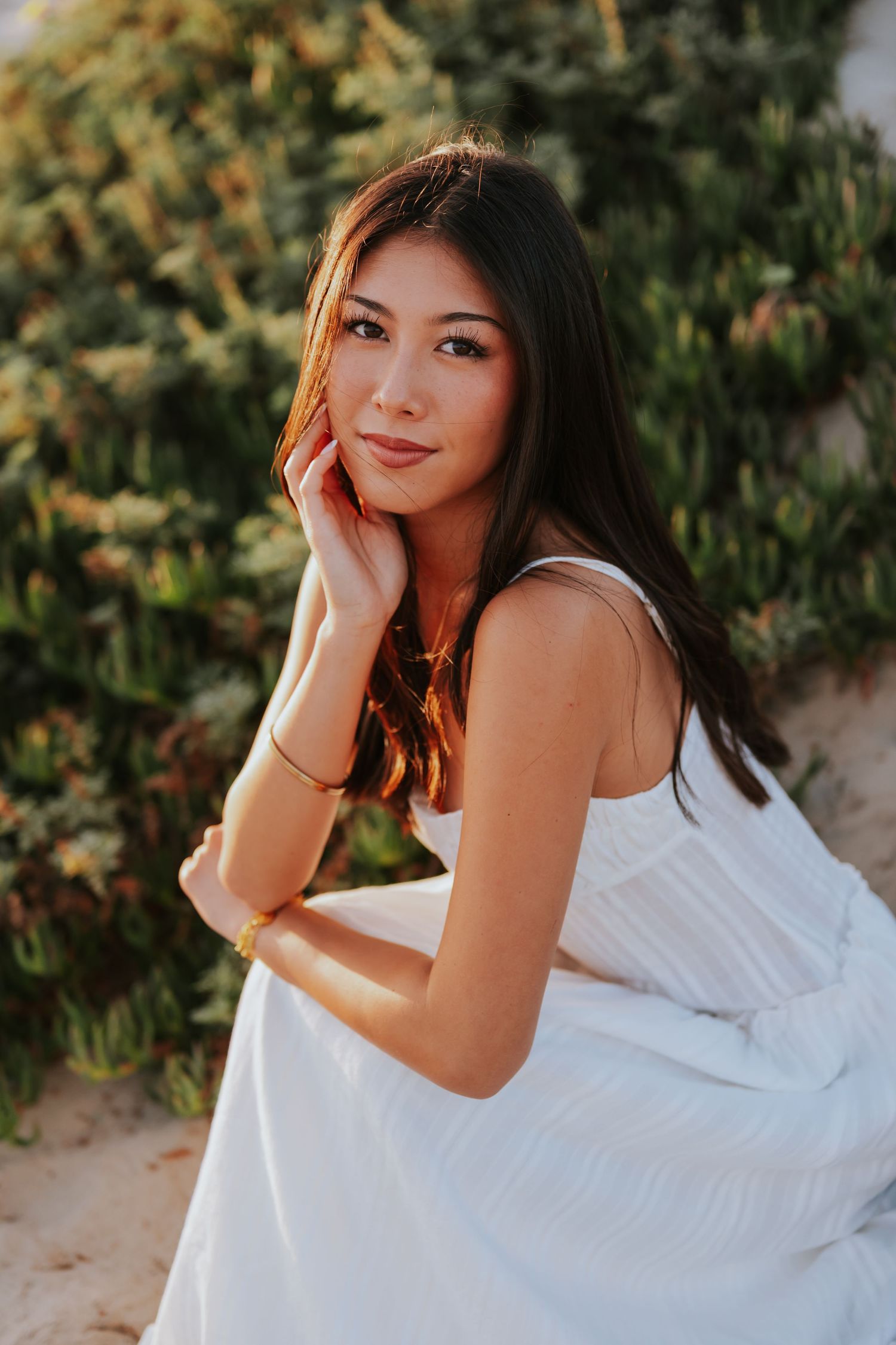 Young woman in white dress sitting outdoors near green bushes, posing gracefully in warm golden light.