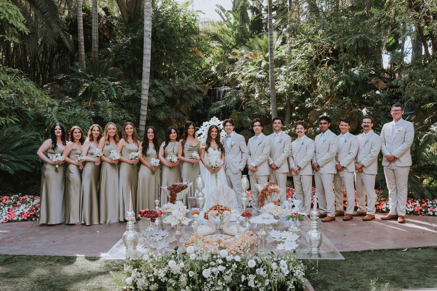 Large wedding party in white attire posed together in lush tropical garden setting.