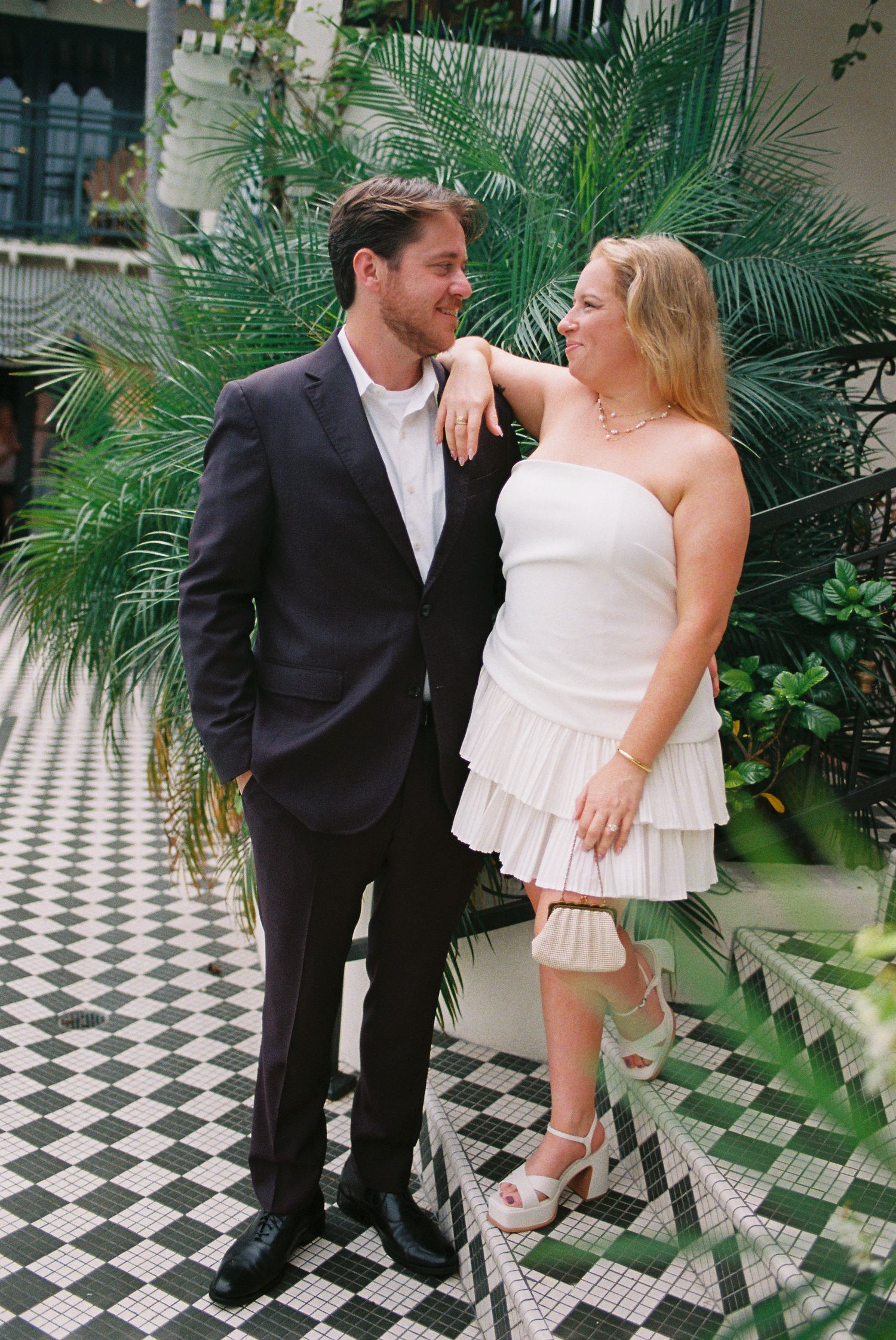 Couple in love posing in tropical courtyard, woman in white ruffled strapless dress, man in dark suit on checkered floor.