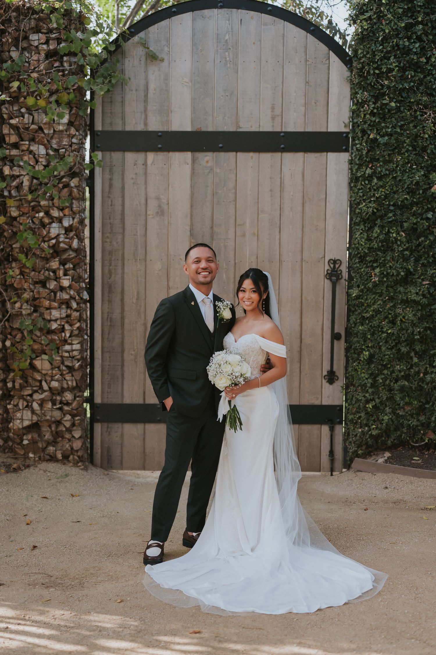 A wedding couple poses in front of a rustic wooden arch door with greenery, the bride wearing a flowing white gown.