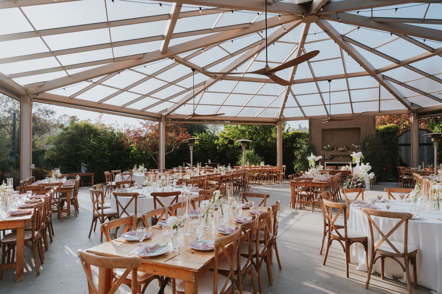 A bright greenhouse-style restaurant interior with wooden tables and chairs arranged under a glass ceiling with natural lighting.