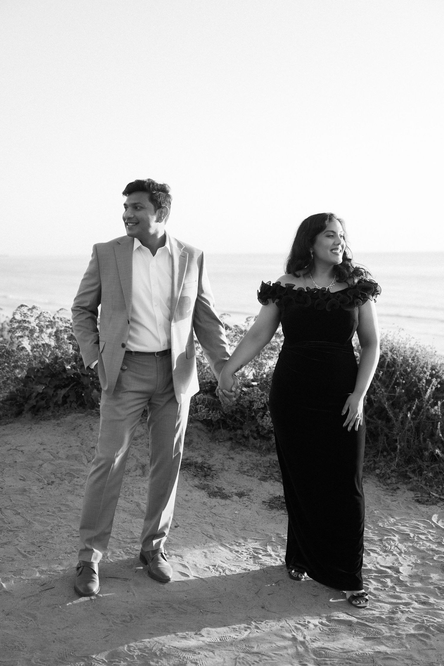Couple holding hands walking on sandy beach path, man in light suit, woman in black off-shoulder gown, black and white photo.
