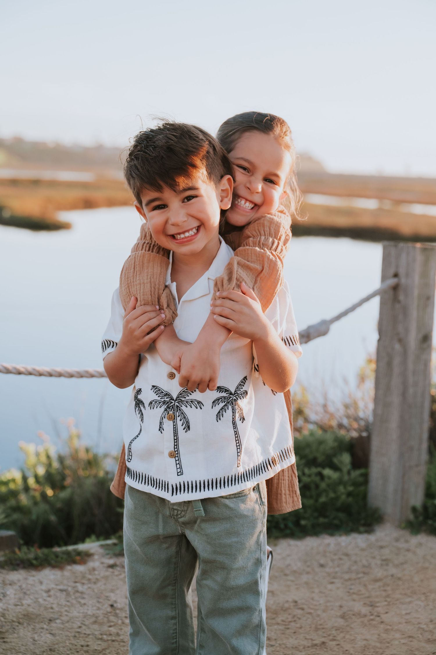 Three candid photos of two children playing and laughing together outdoors near a wooden fence at sunset.