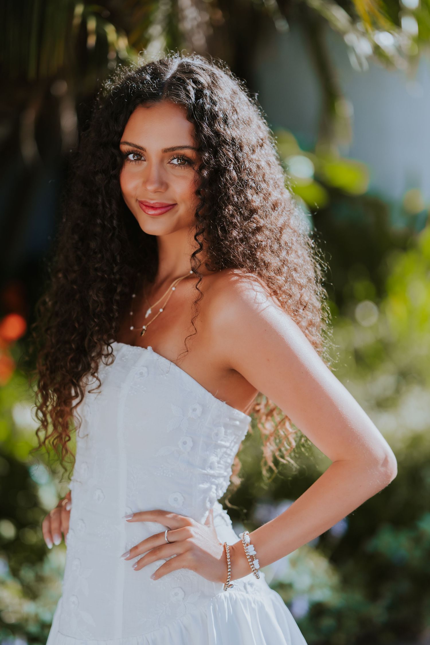 A bride in a white strapless wedding dress with curly hair poses outdoors in natural sunlight.