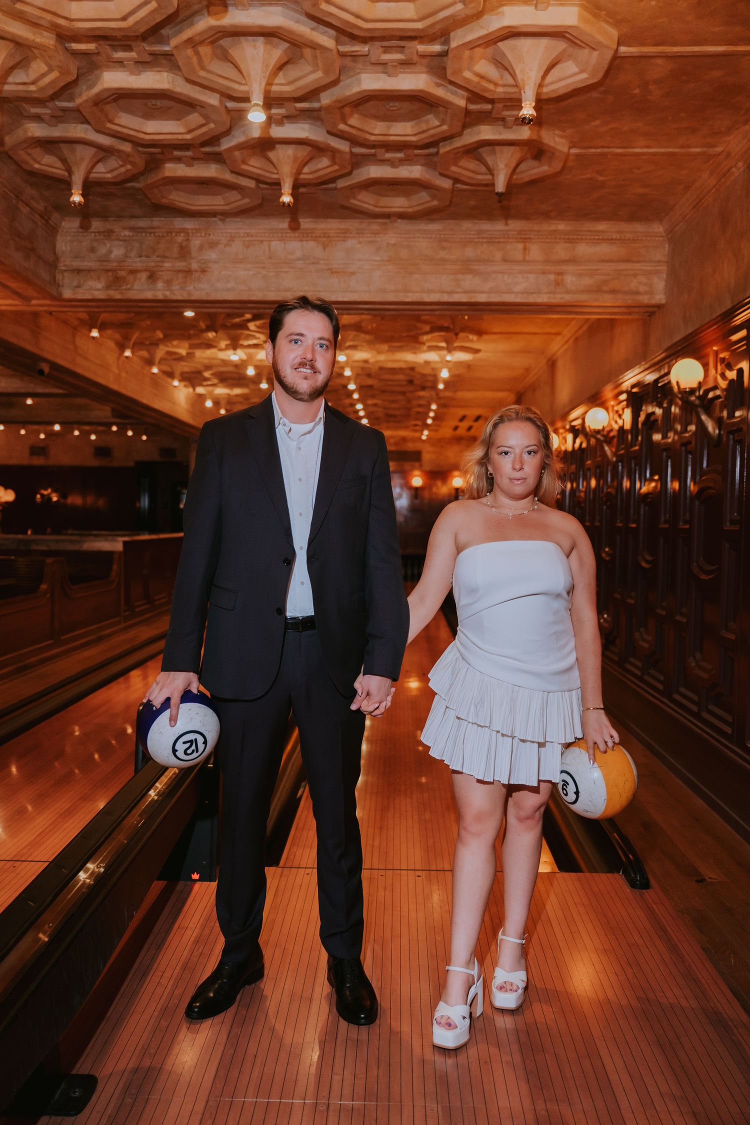 Bride and groom in white mini dress and black suit holding bowling balls in a vintage bowling alley.