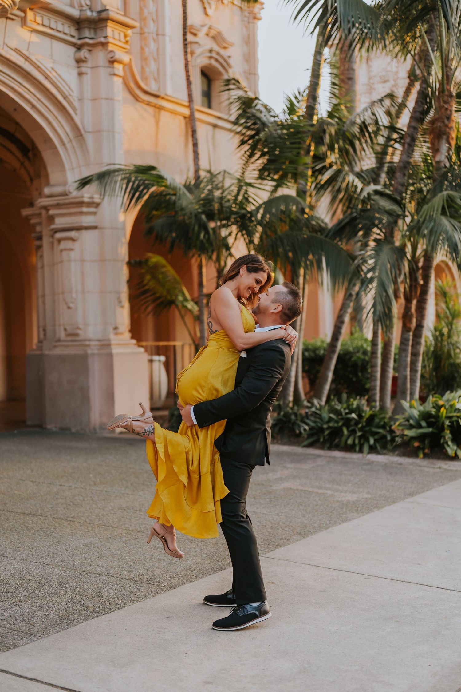 A couple in yellow and black outfits share a romantic moment in front of a grand archway with palm trees at sunset.