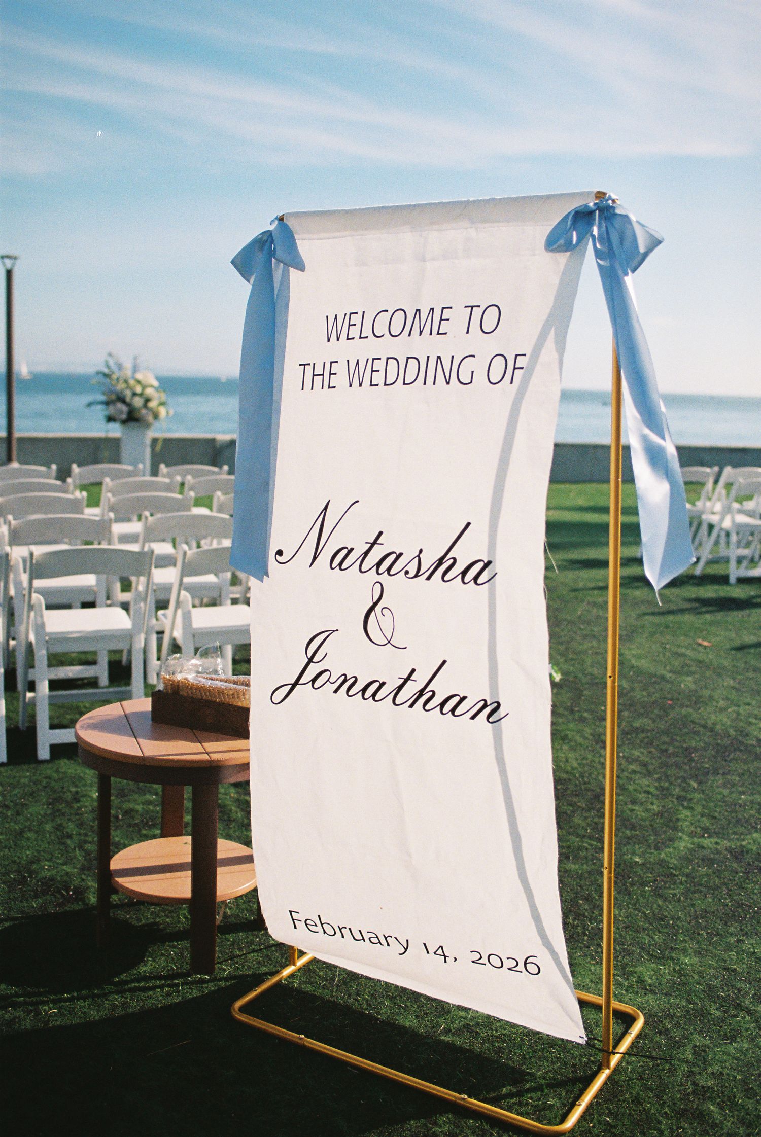 Outdoor wedding welcome sign for Natasha and Jonathan, February 14 2026, displayed on gold stand with blue ribbon bows.
