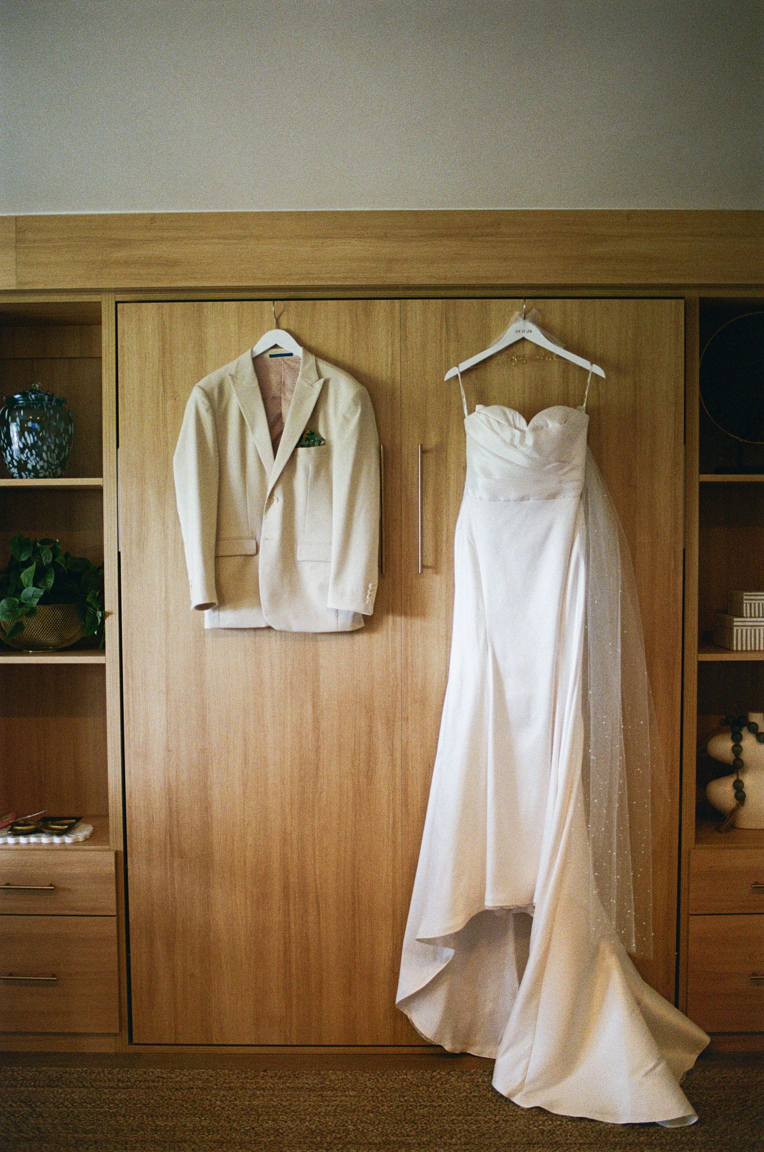 Wedding dress and groom suit hanging on wooden wardrobe before ceremony.