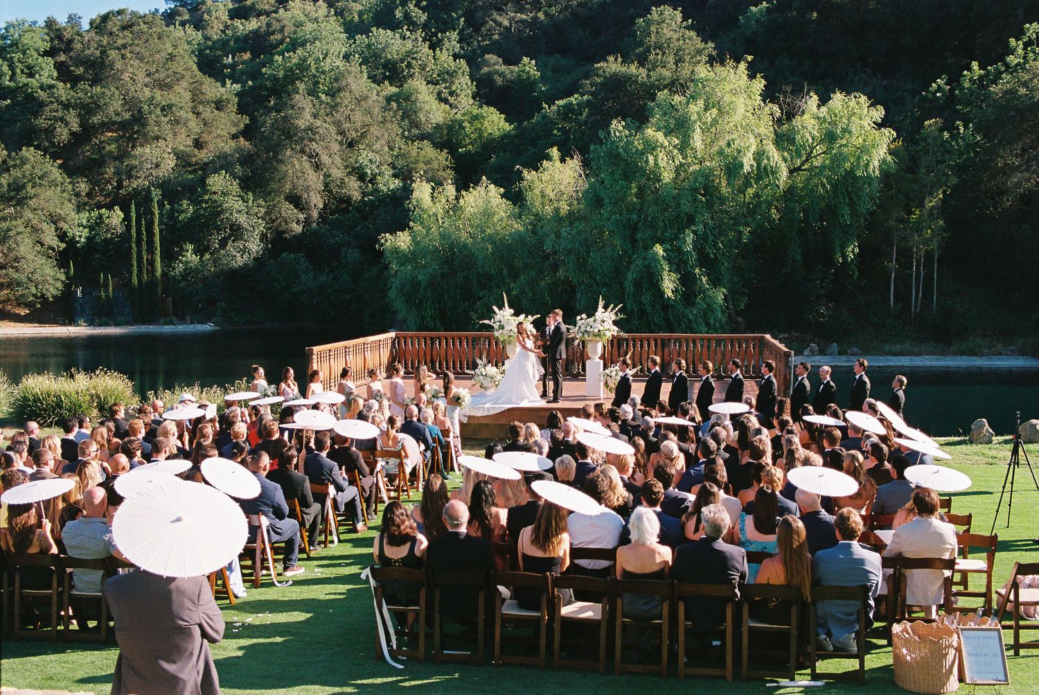 Outdoor wedding ceremony on lush green lawn with guests seated in rows, white umbrellas, and scenic tree-lined hills backdrop.