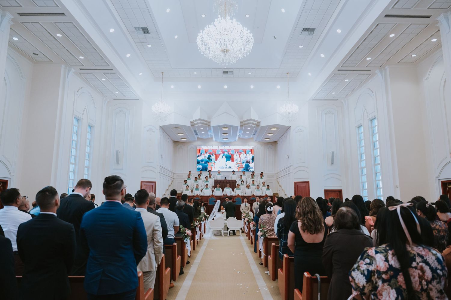 Elegant wedding ceremony in a bright white ballroom with crystal chandeliers and rows of wooden pews.
