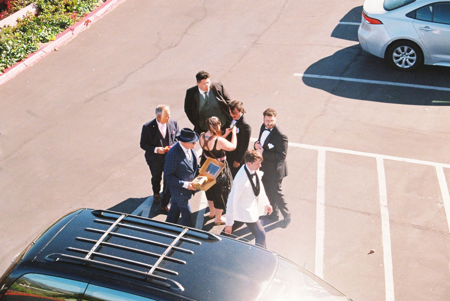 Wedding guests gathered in a parking lot area near parked cars during an outdoor celebration.