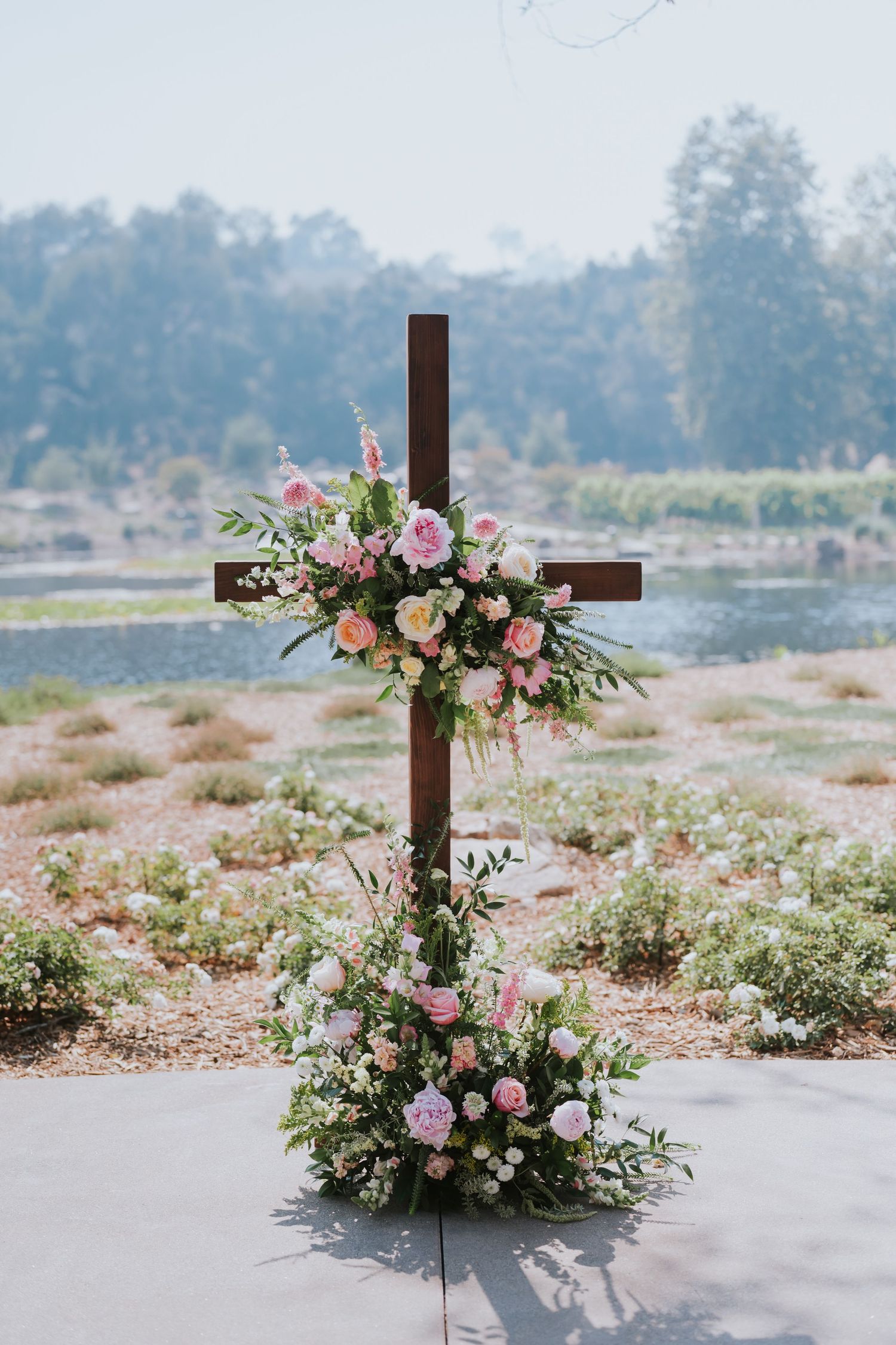 Wooden cross decorated with pink and white floral arrangements at an outdoor wedding ceremony.