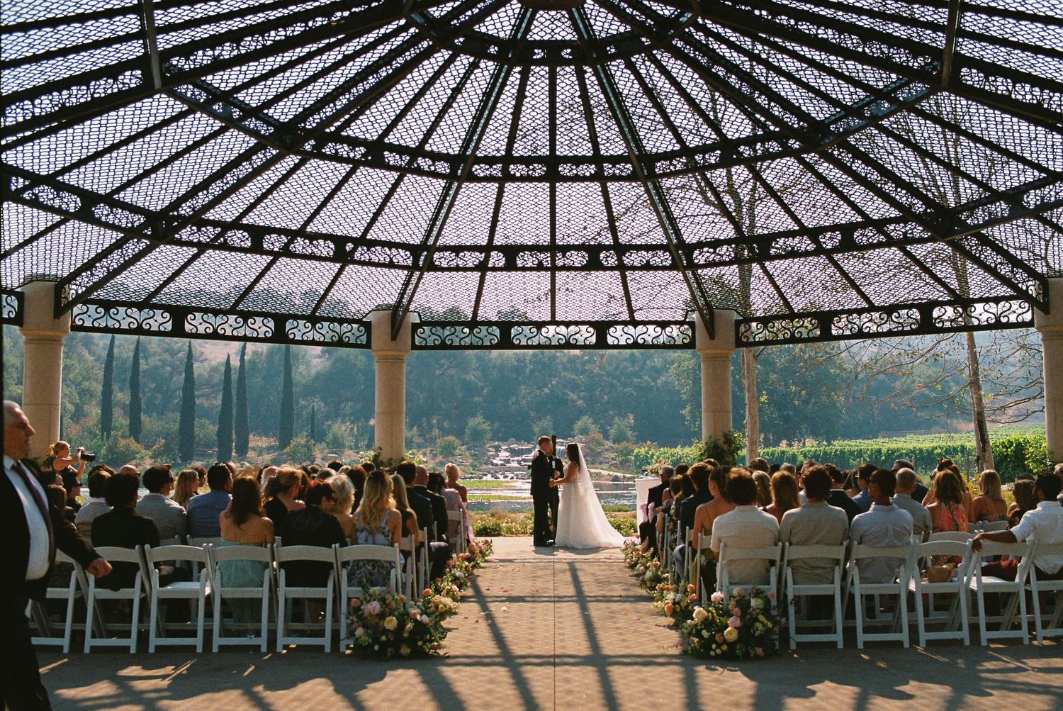 Elegant outdoor wedding ceremony under ornate iron gazebo with guests seated in white chairs surrounded by lush gardens.