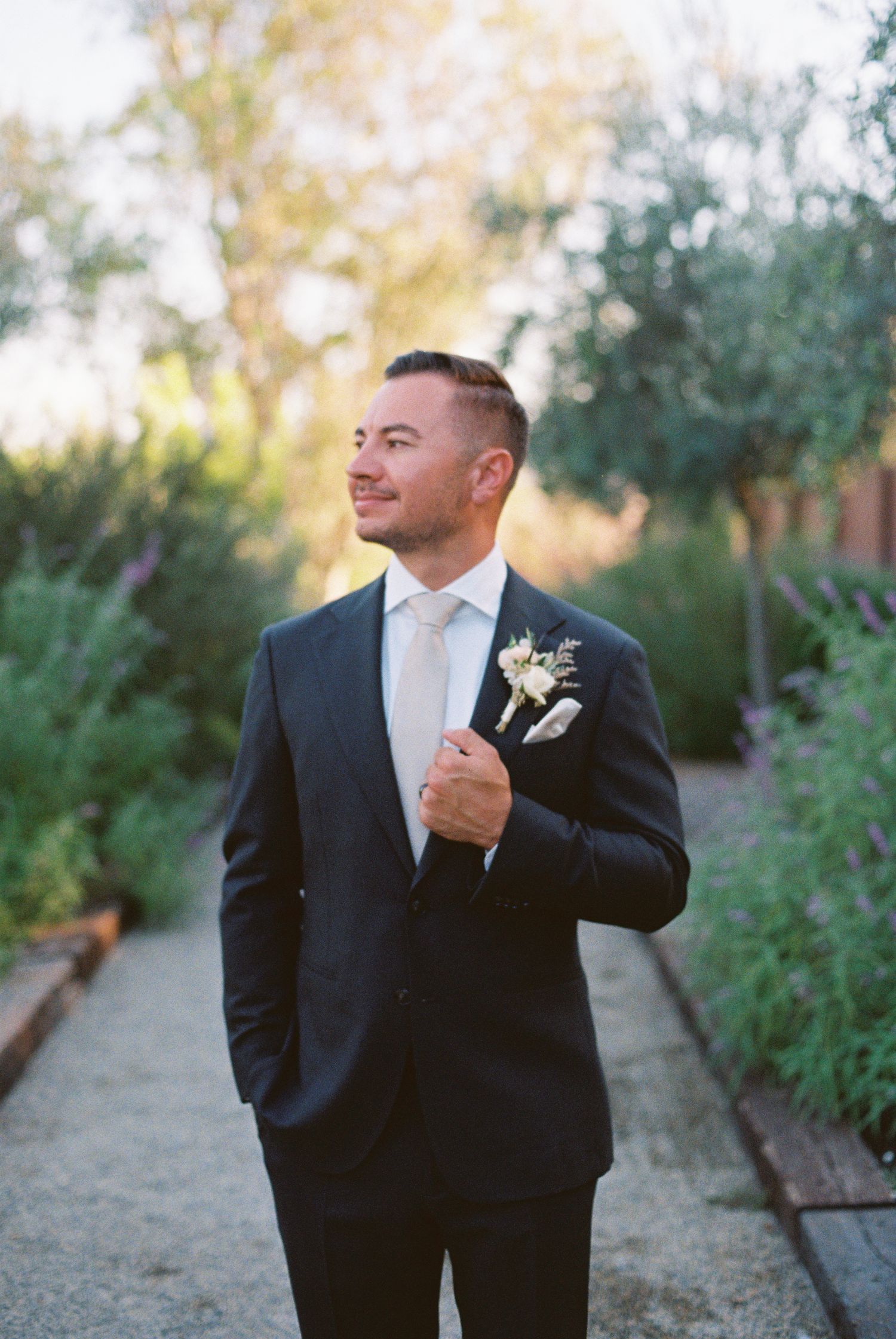 Groom in dark suit with white boutonniere and tie stands on garden path, smiling, with lush greenery in background.