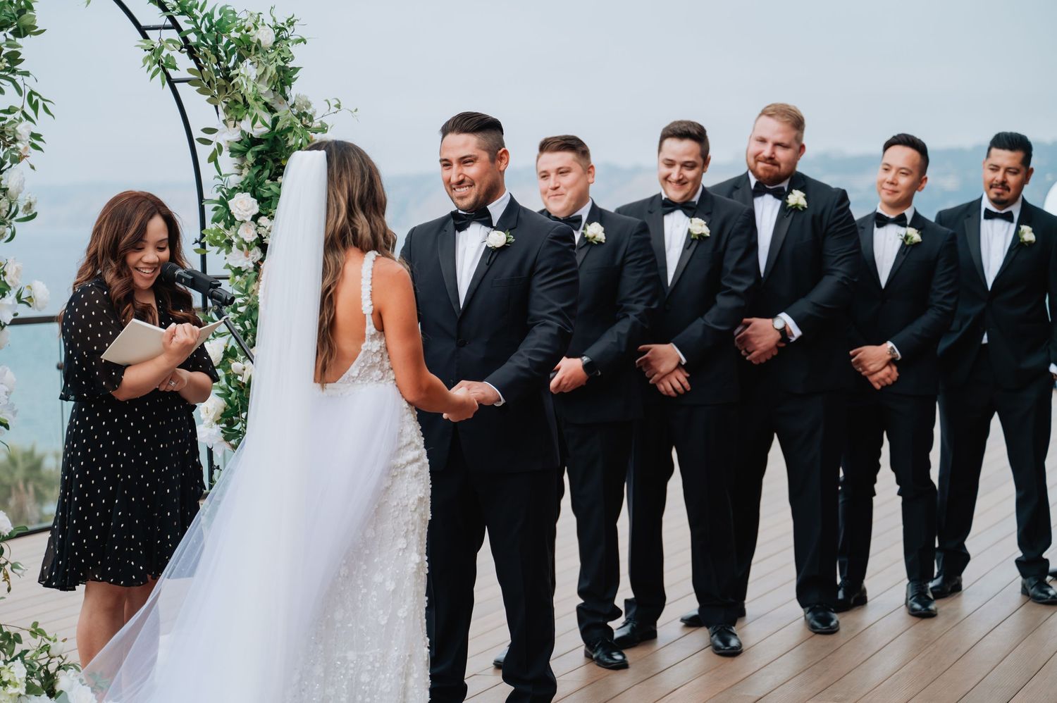 Wedding party in formal attire poses together on oceanfront deck.