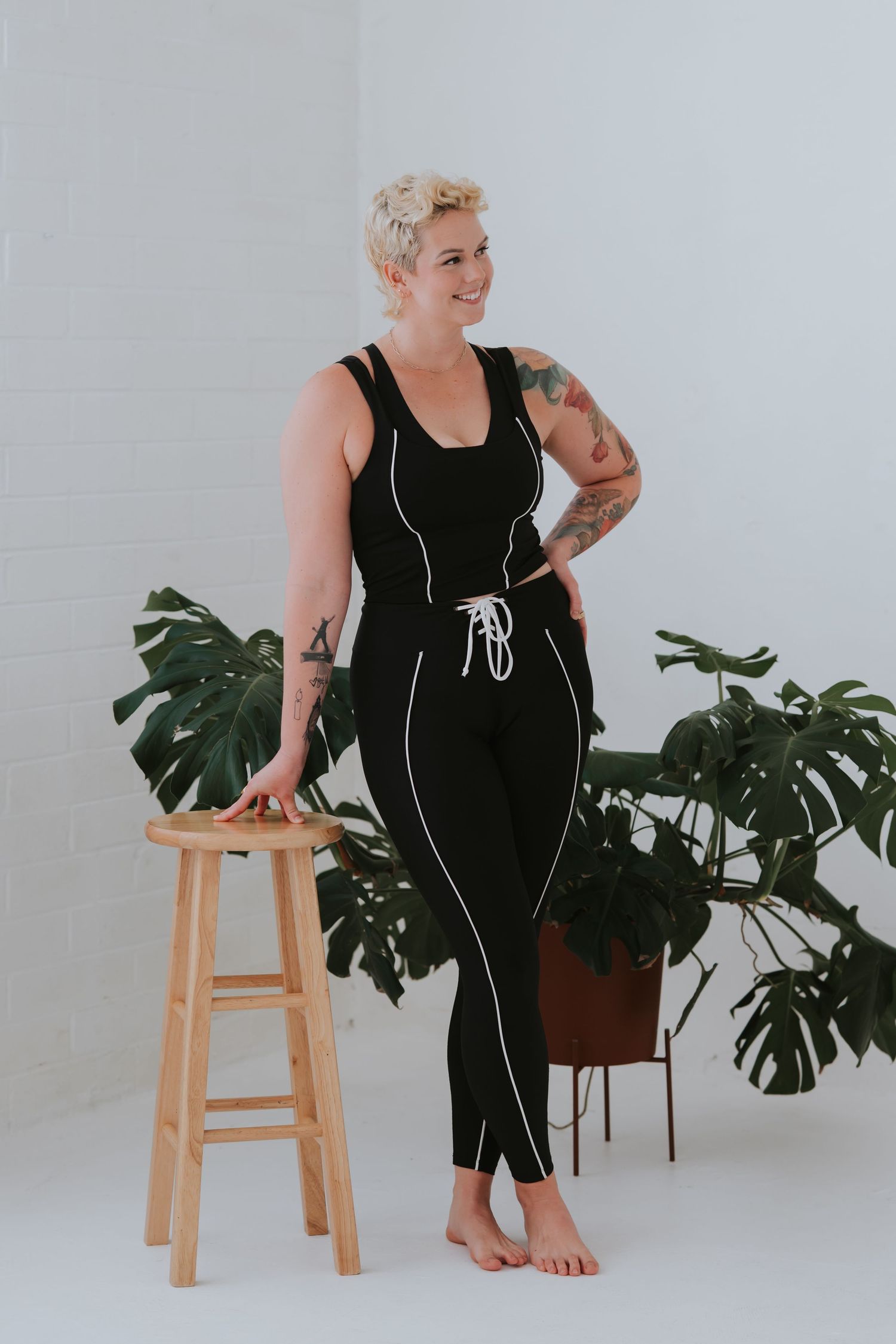 Person in black athleisure outfit sits on wooden stool next to large monstera plant against white backdrop.
