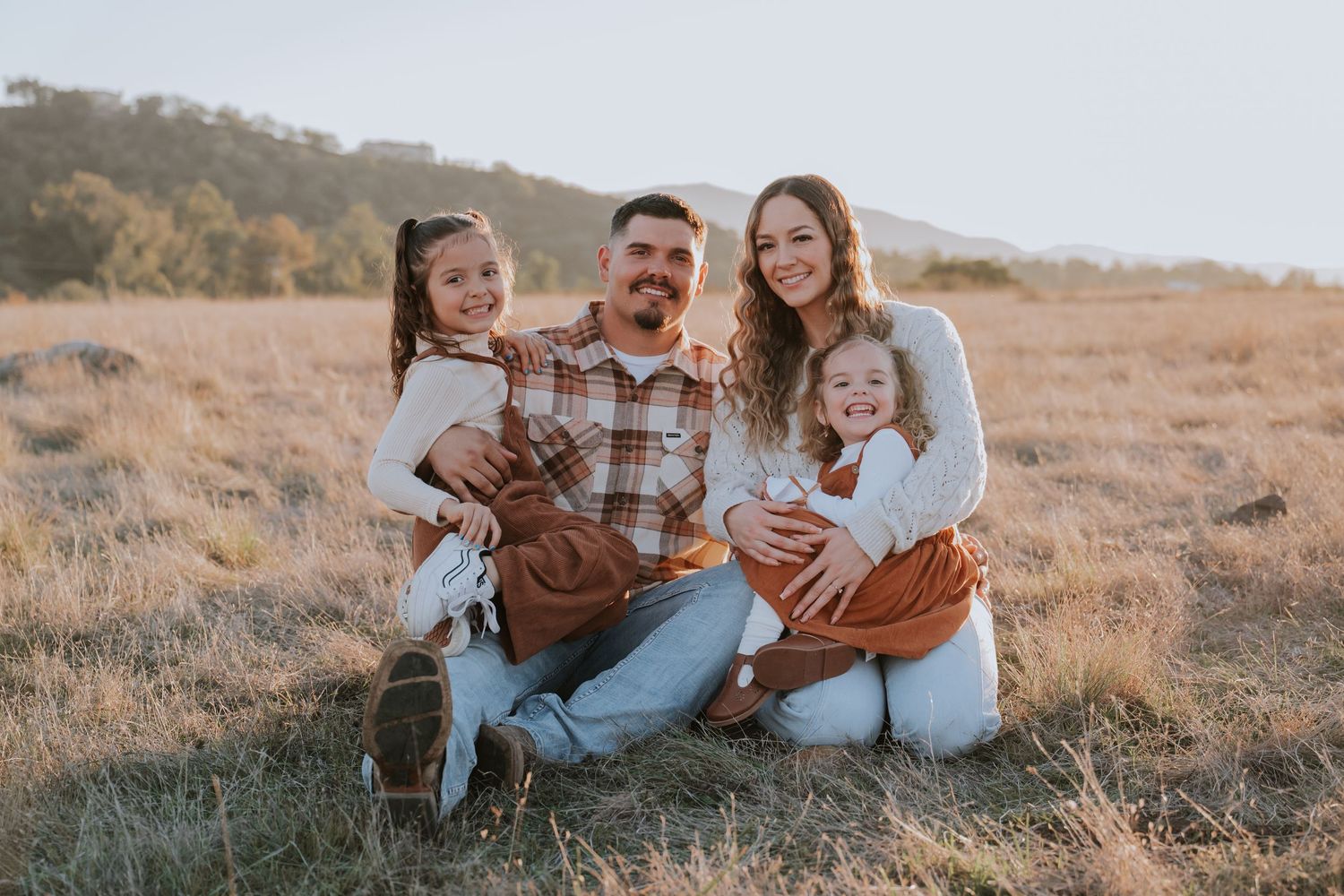 Happy family of four sitting in a golden field, parents smiling with two young daughters on a sunny day.