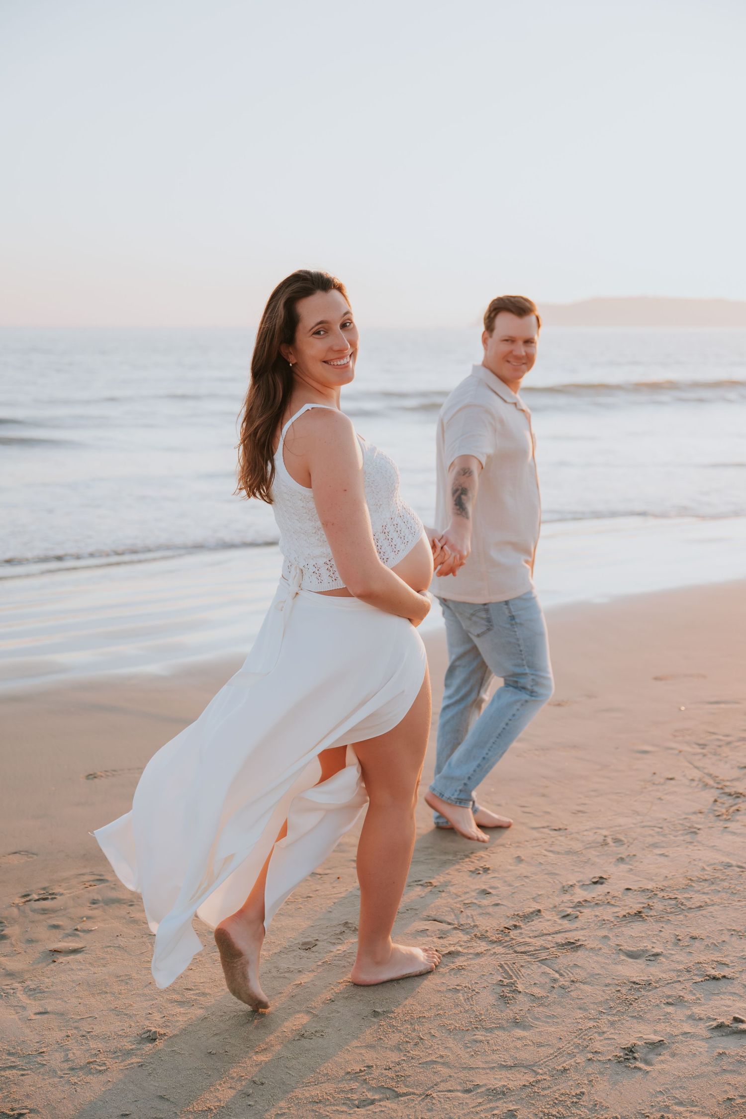 Couple holding hands walking on beach at sunset, woman in white flowing dress smiling back at camera.