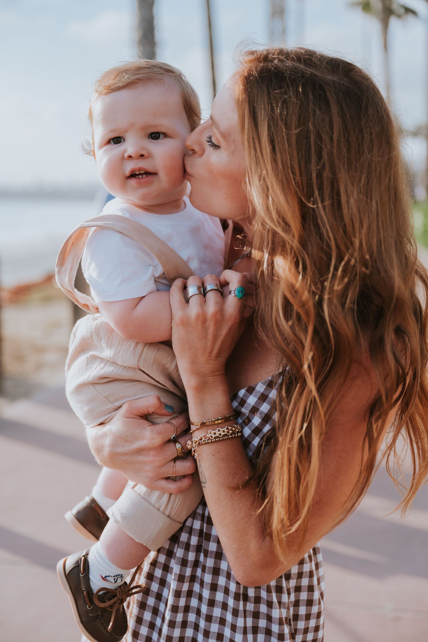 A woman with long hair kisses a baby on the cheek while holding him outdoors near a beach.
