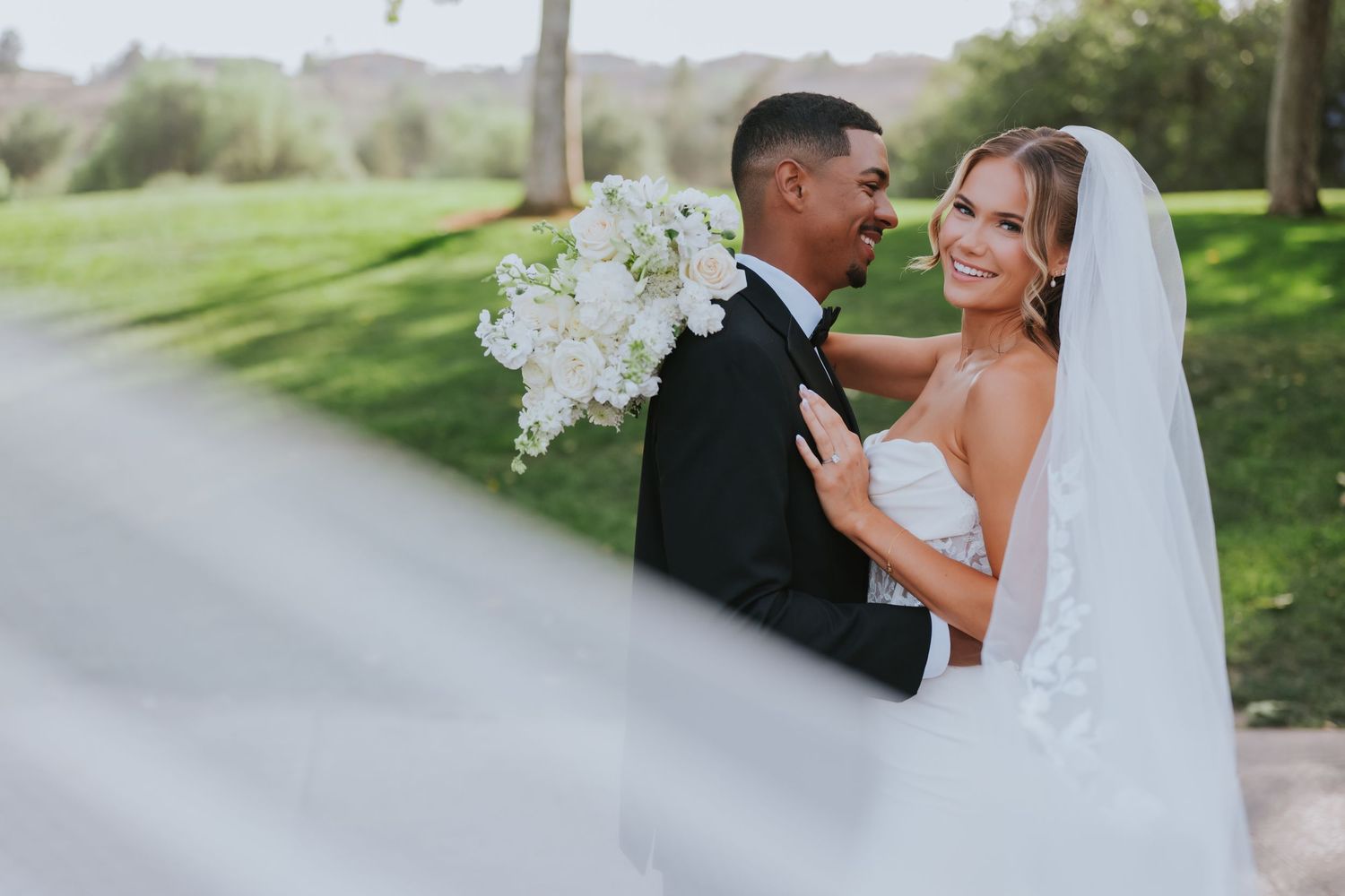 Bride and groom embrace outdoors, she holds white bouquet, veil flows, lush green background, elegant wedding attire.