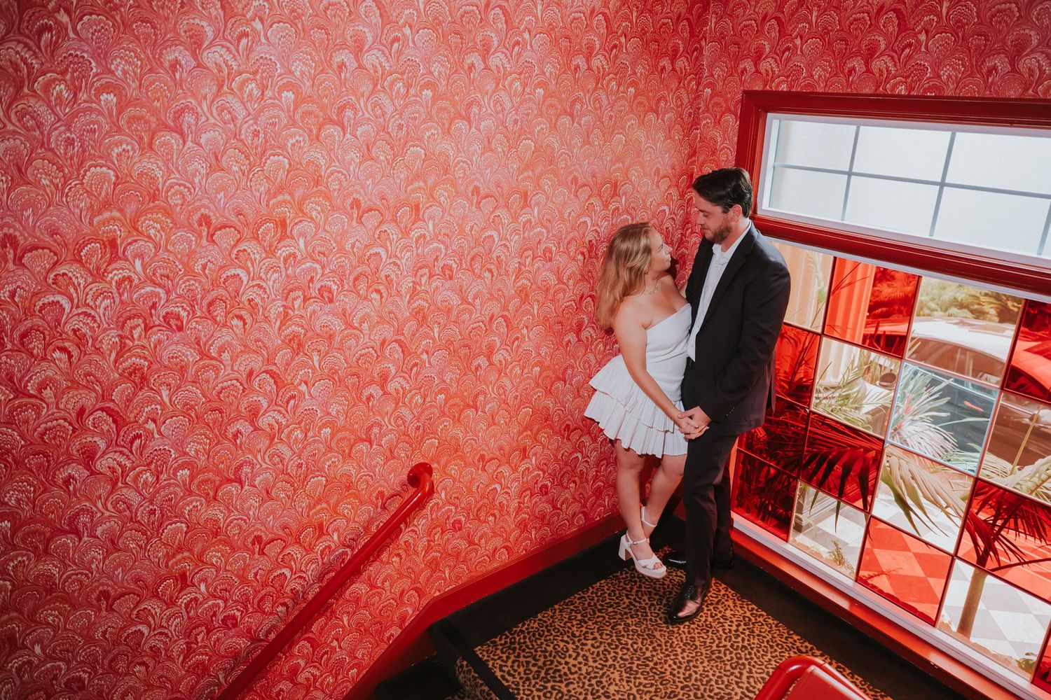 Couple sharing a romantic moment on a staircase with vibrant red patterned wallpaper in an elegant venue.