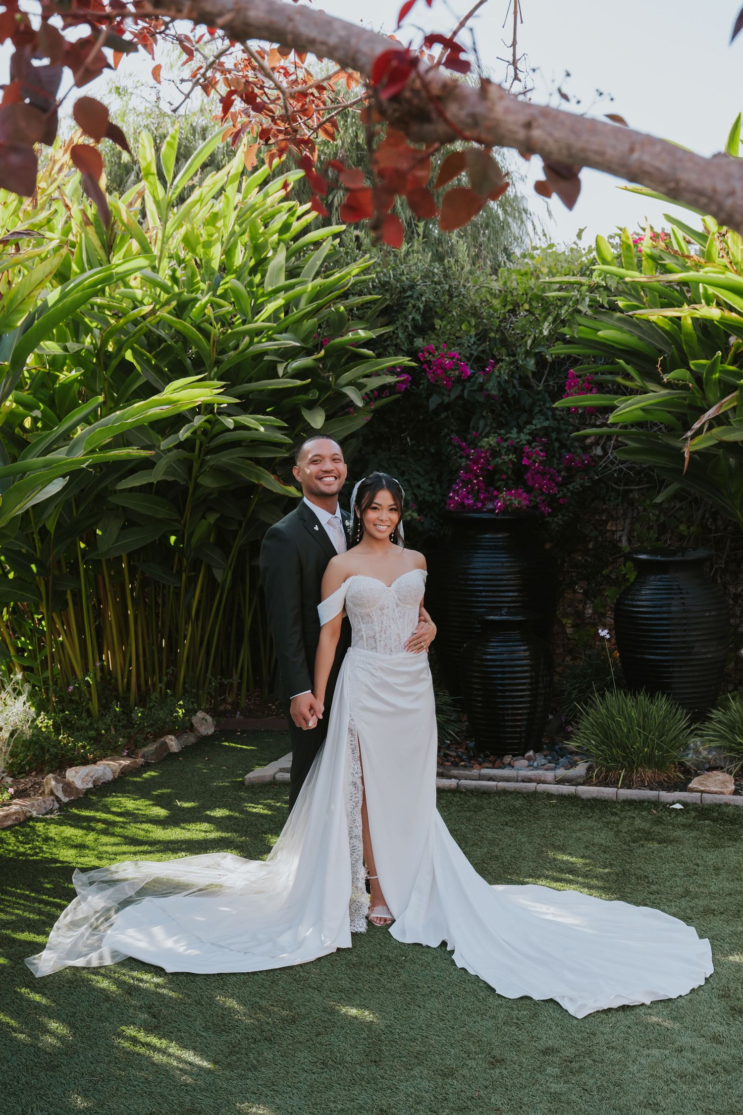 Wedding couple poses together on manicured lawn with tropical garden backdrop and bamboo plants.