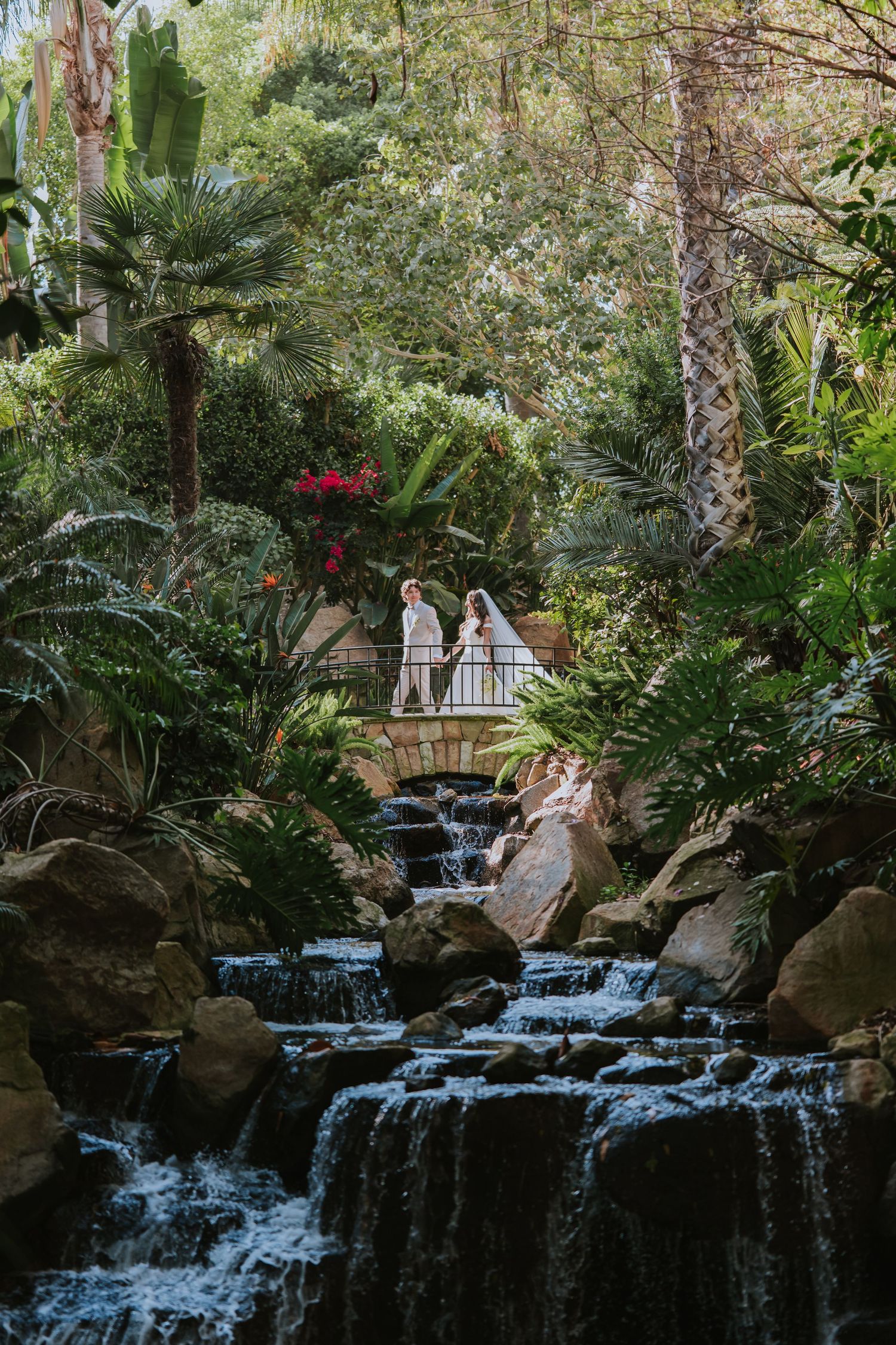 A scenic waterfall cascades down rocks in a lush tropical garden setting with a viewing deck above.