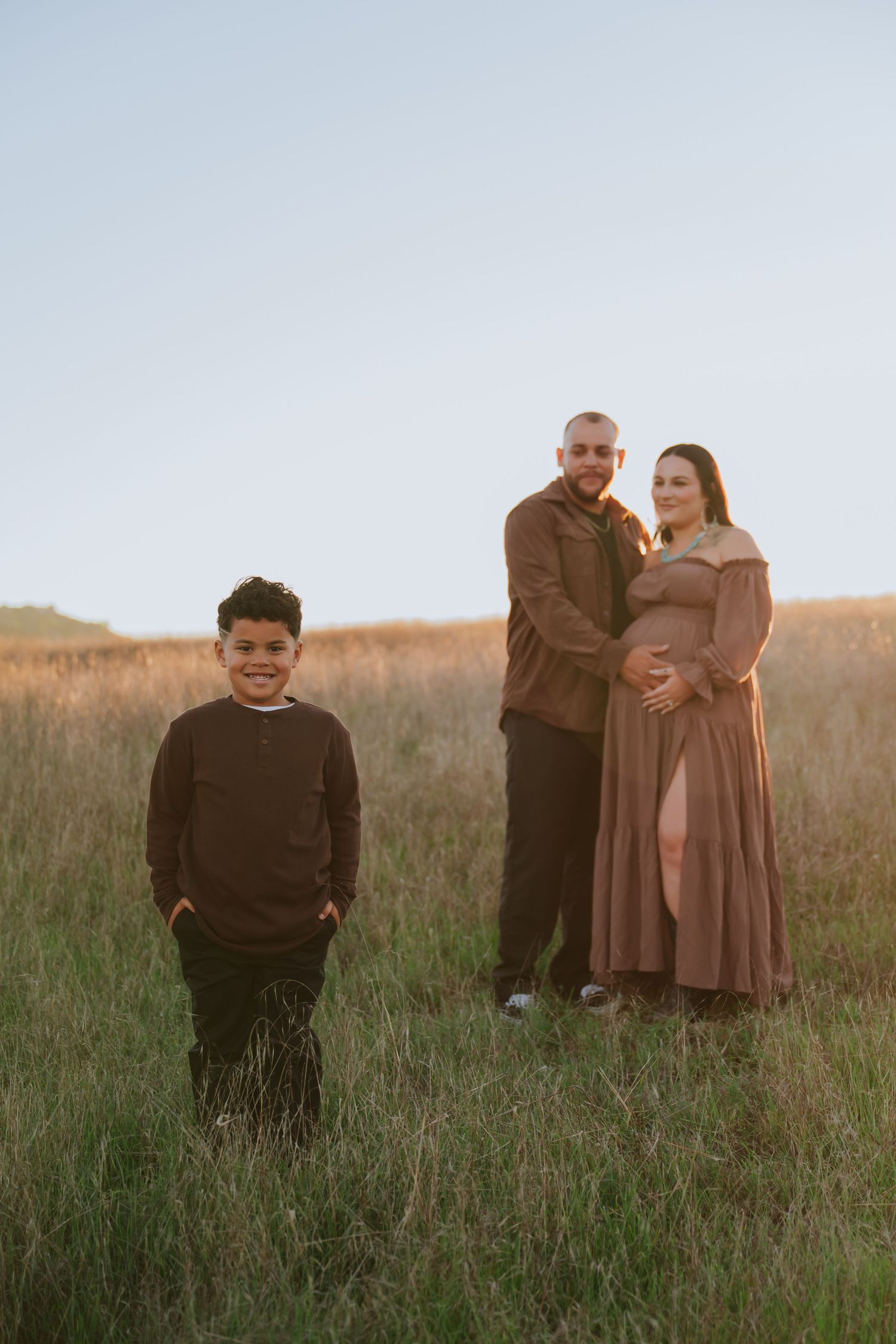 Maternity photo of pregnant woman with partner and young boy in golden field at sunset.