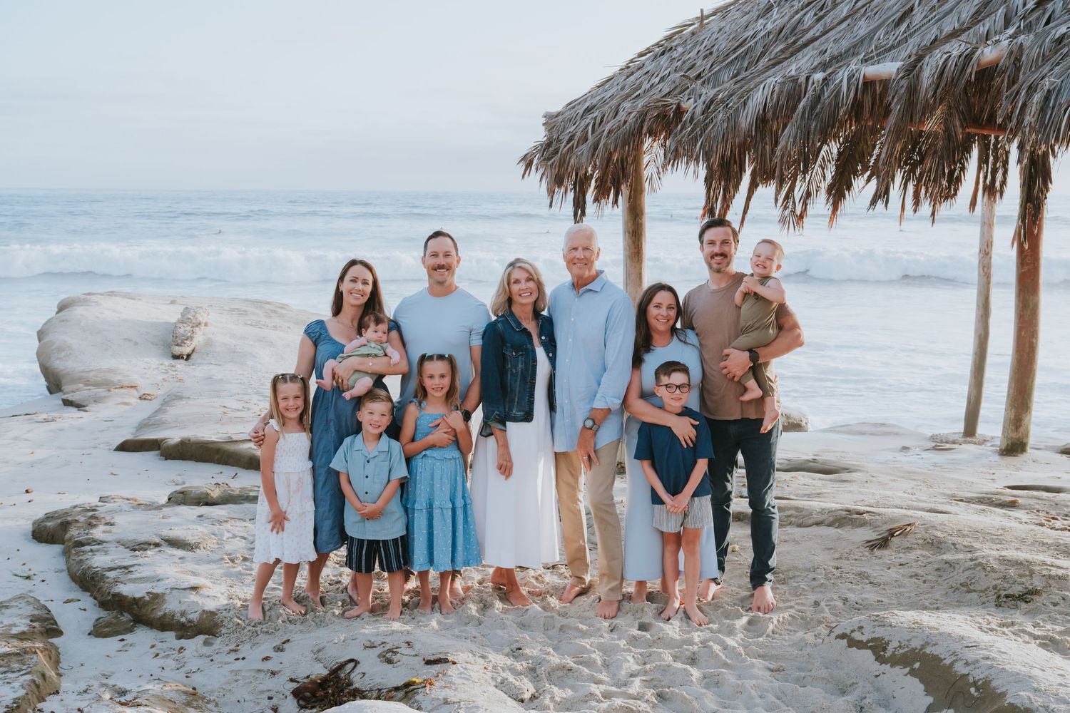 Large family posing together on a rocky beach near a thatched palapa, with ocean waves in the background.